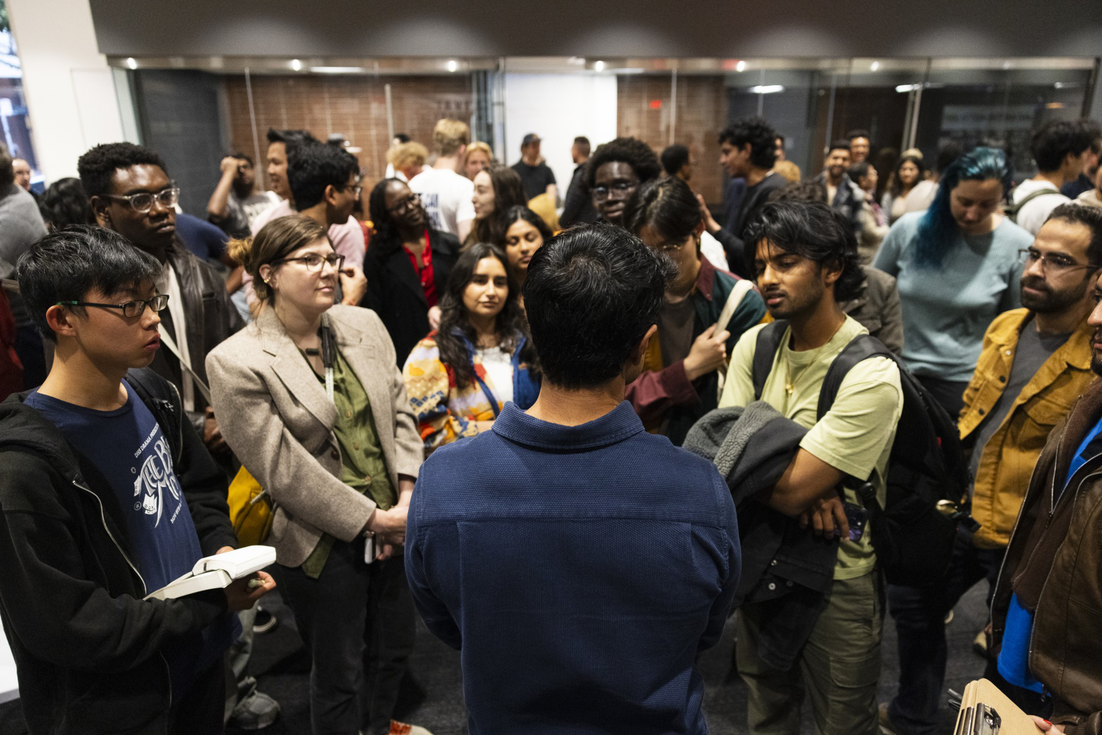 A large group of diverse people gathers closely indoors, attentively listening to a man in a blue shirt who has his back to the camera.