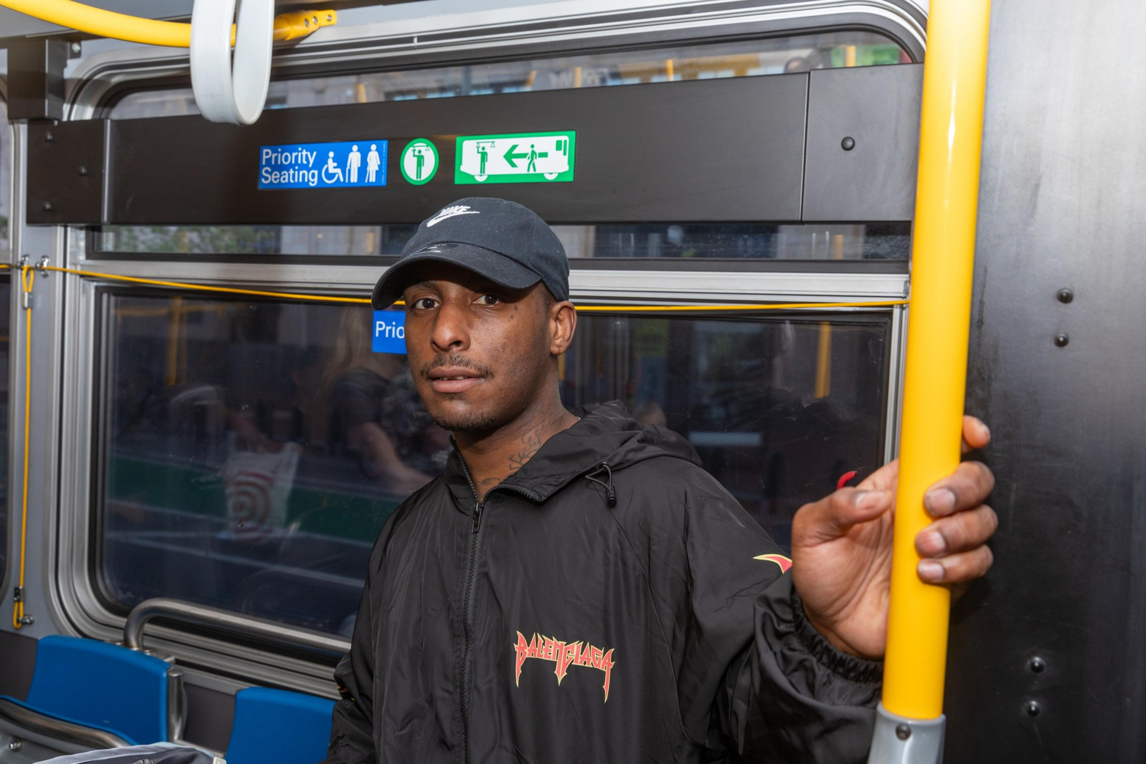 A man wearing a black jacket and cap is standing inside a bus, holding a yellow pole. There are signs above for priority seating and emergency exits.