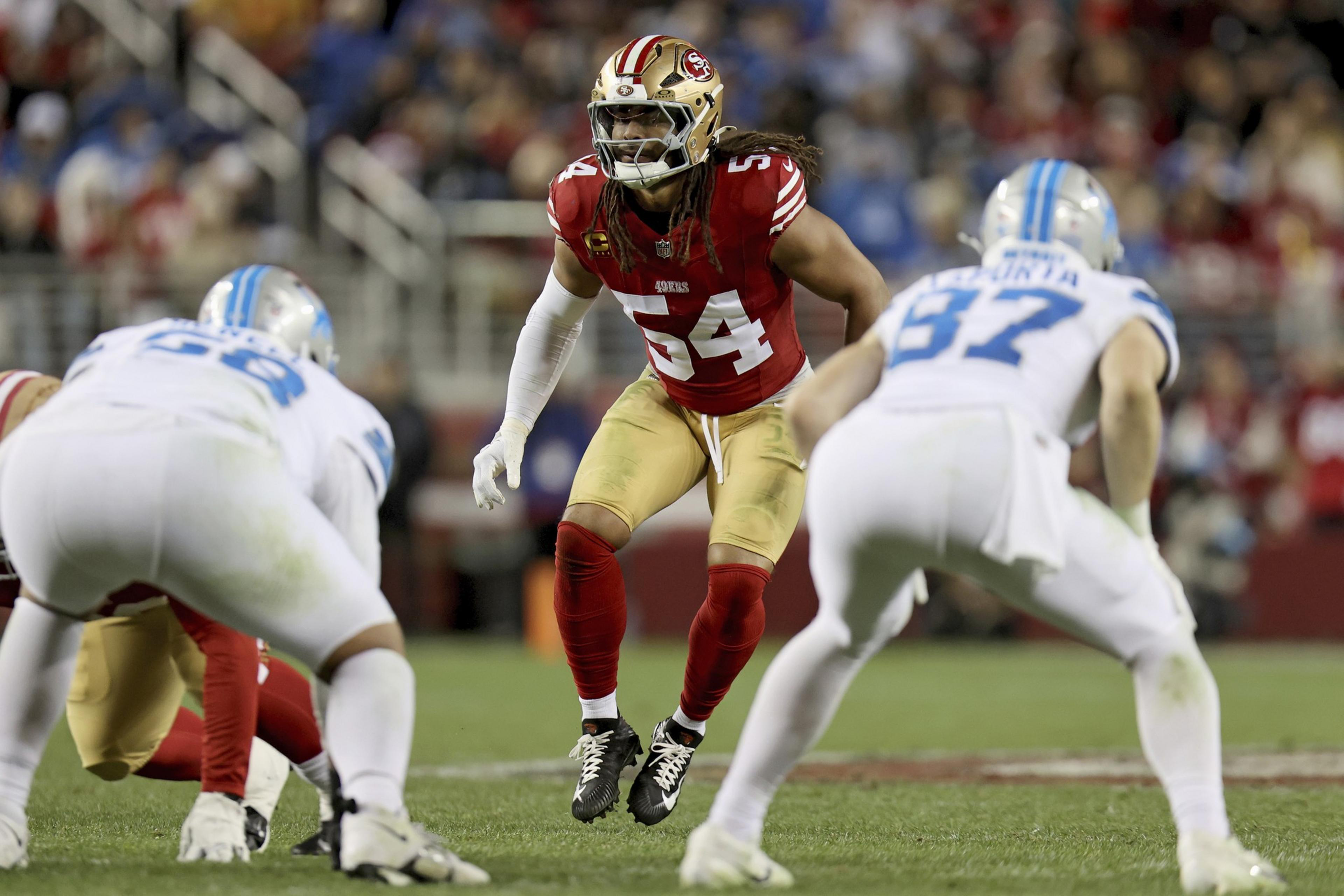 A football player in a red and gold jersey is poised and ready on a field, surrounded by players in white and blue uniforms, during a game.