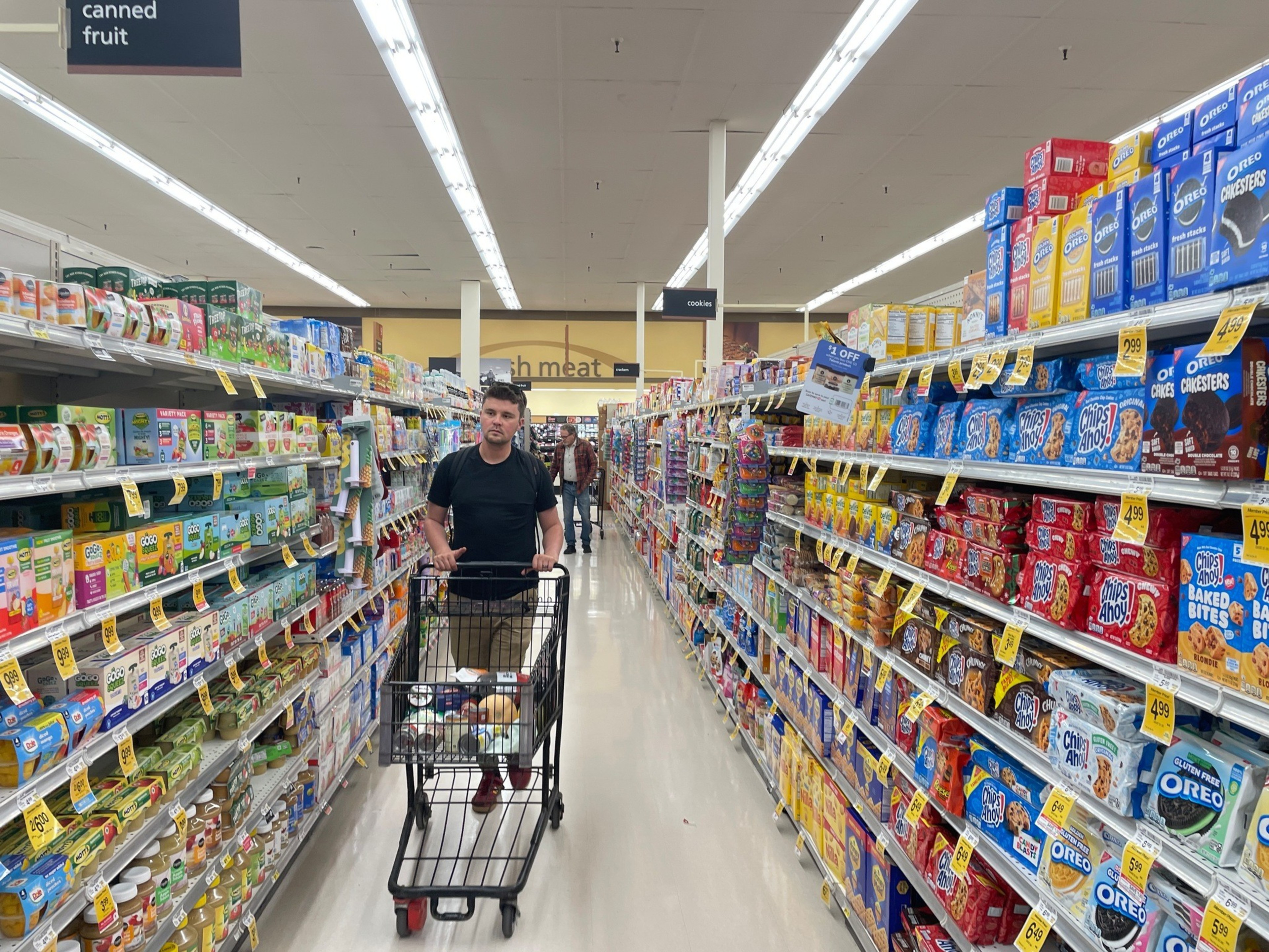 A man pushes a shopping cart down a grocery aisle with canned fruit on one side and various cookie brands on the other.