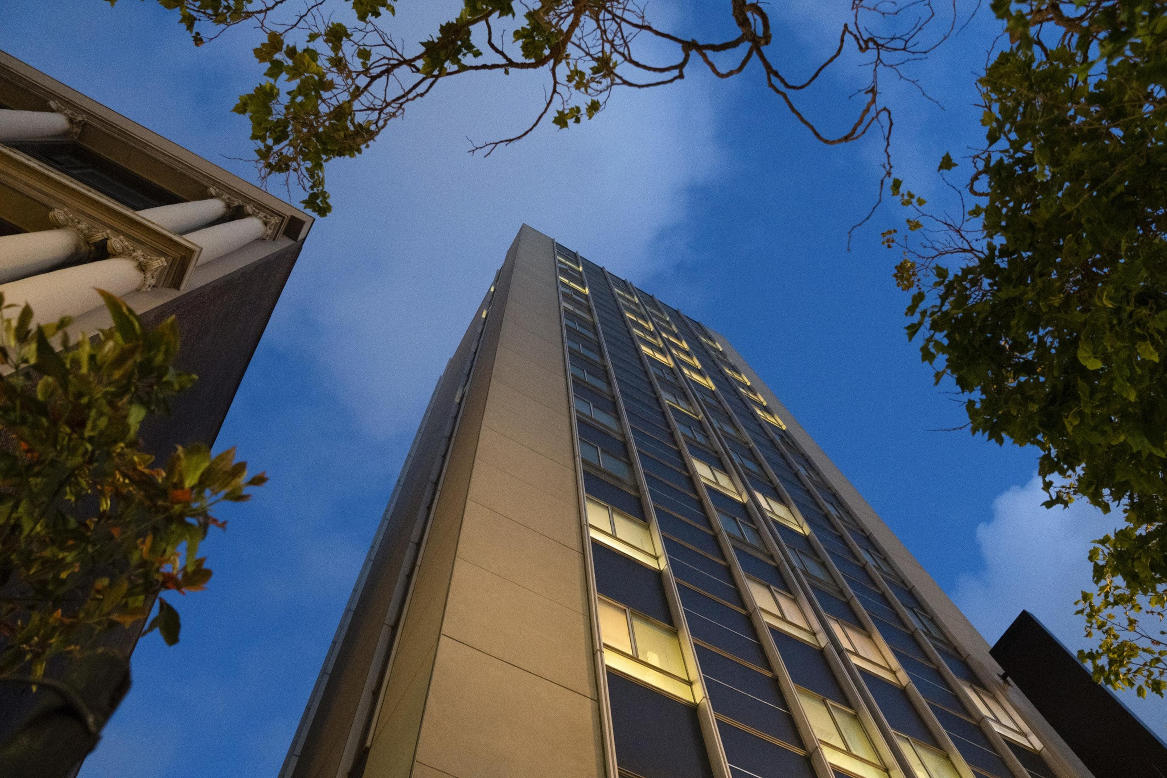 A tall, modern building is seen from below against a twilight sky. Trees frame the image, and the building has many lit windows, giving a sense of urban vibrancy.