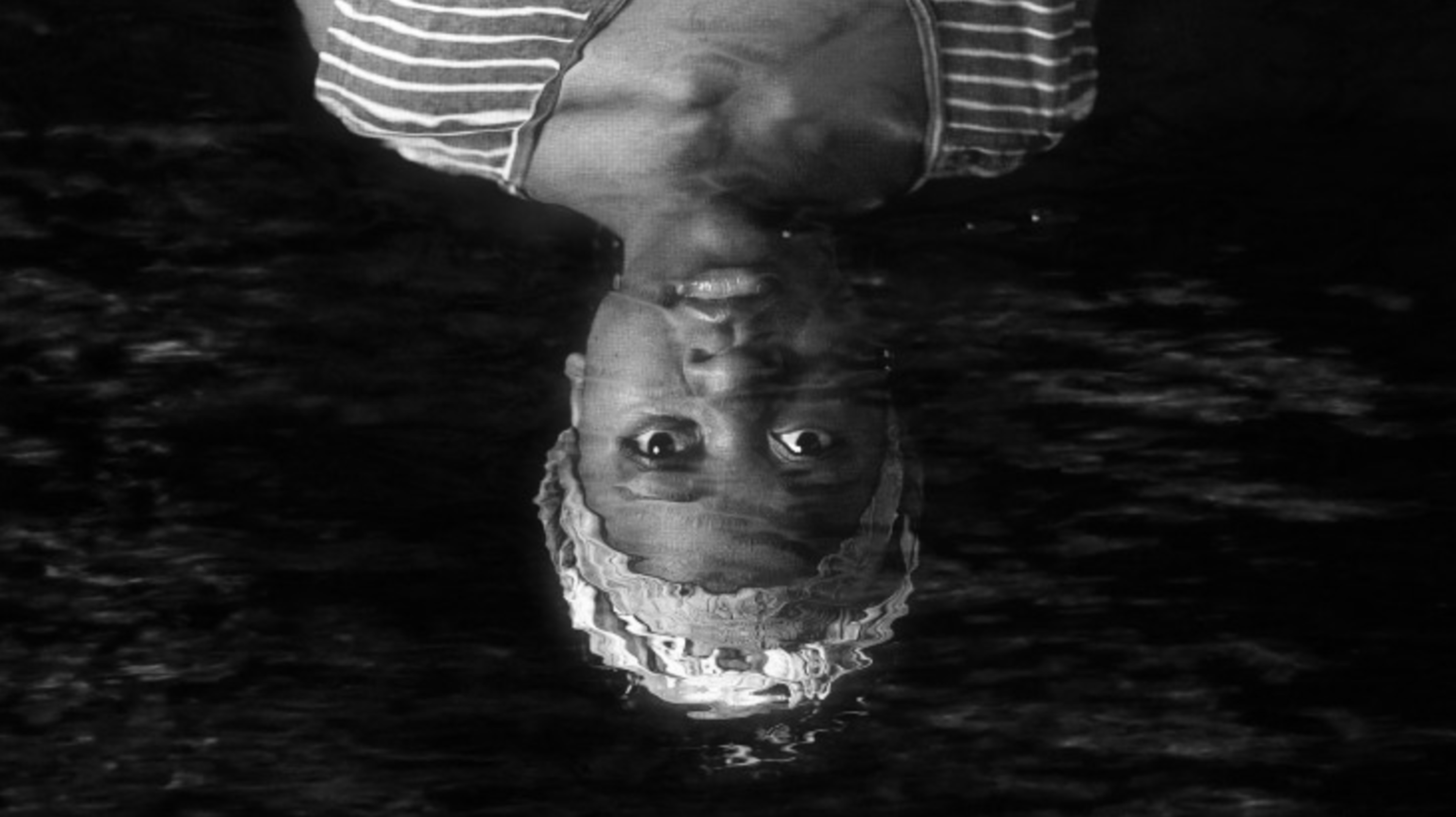 An upside-down black-and-white close-up of a person wearing a striped shirt and a head wrap, reflected in rippling water.