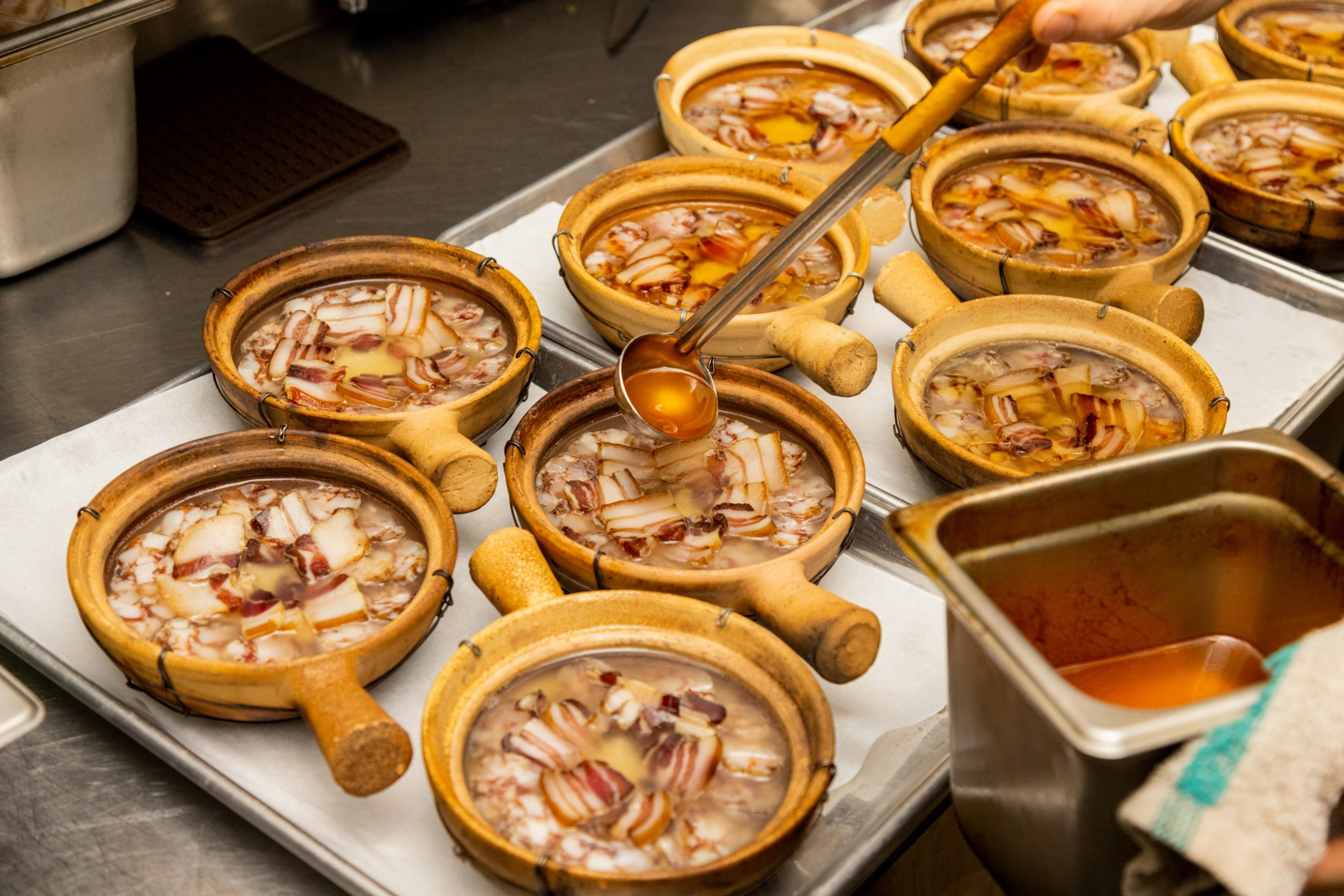 Multiple clay pots with soup and bacon, a hand ladling broth into one.