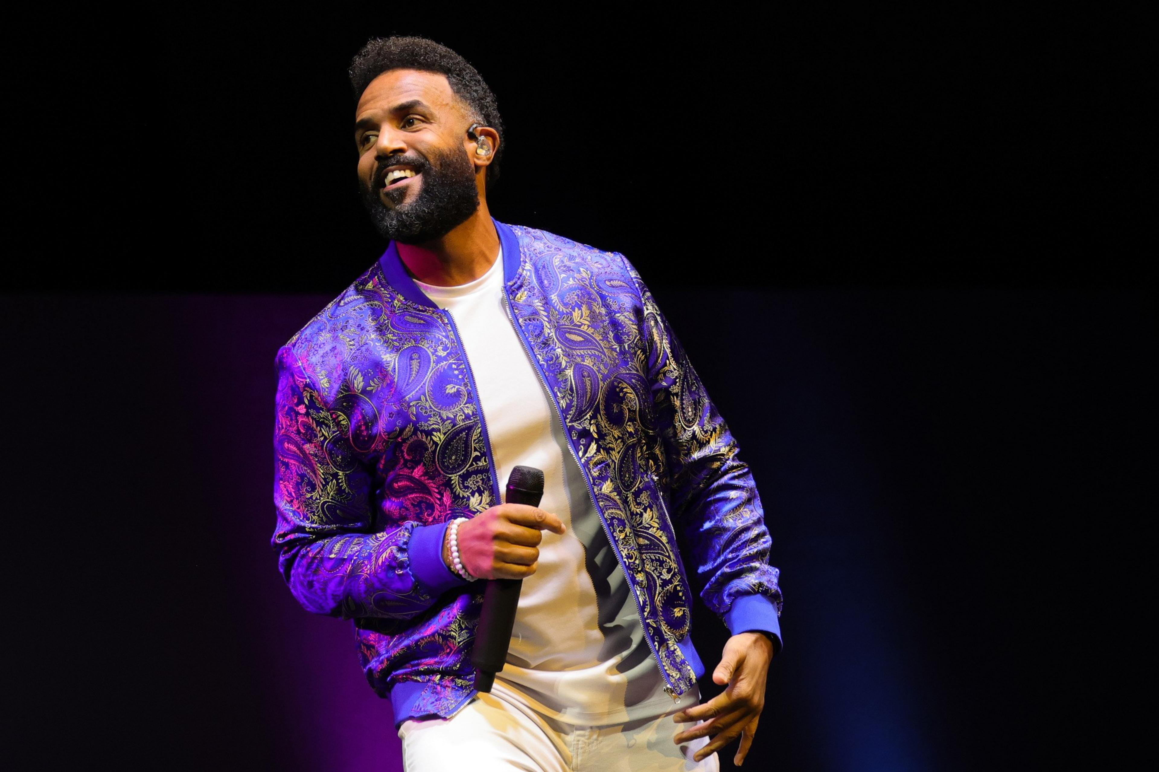 A man on stage with a mic, smiling, wearing a vibrant blue patterned jacket.
