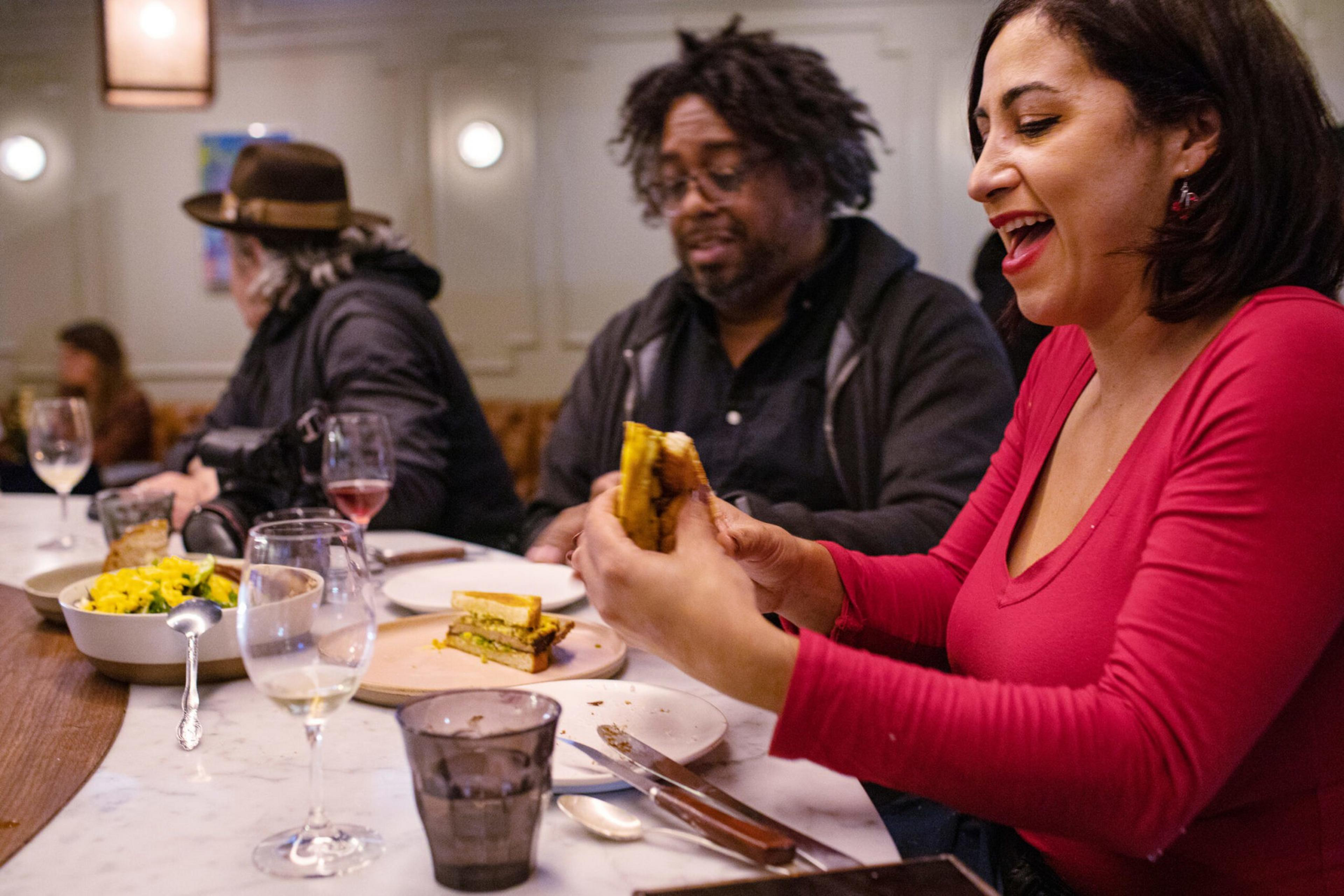 A woman in a red top smiles while holding a sandwich, sitting at a table with two men, plates, glasses, and food in a warmly lit room.