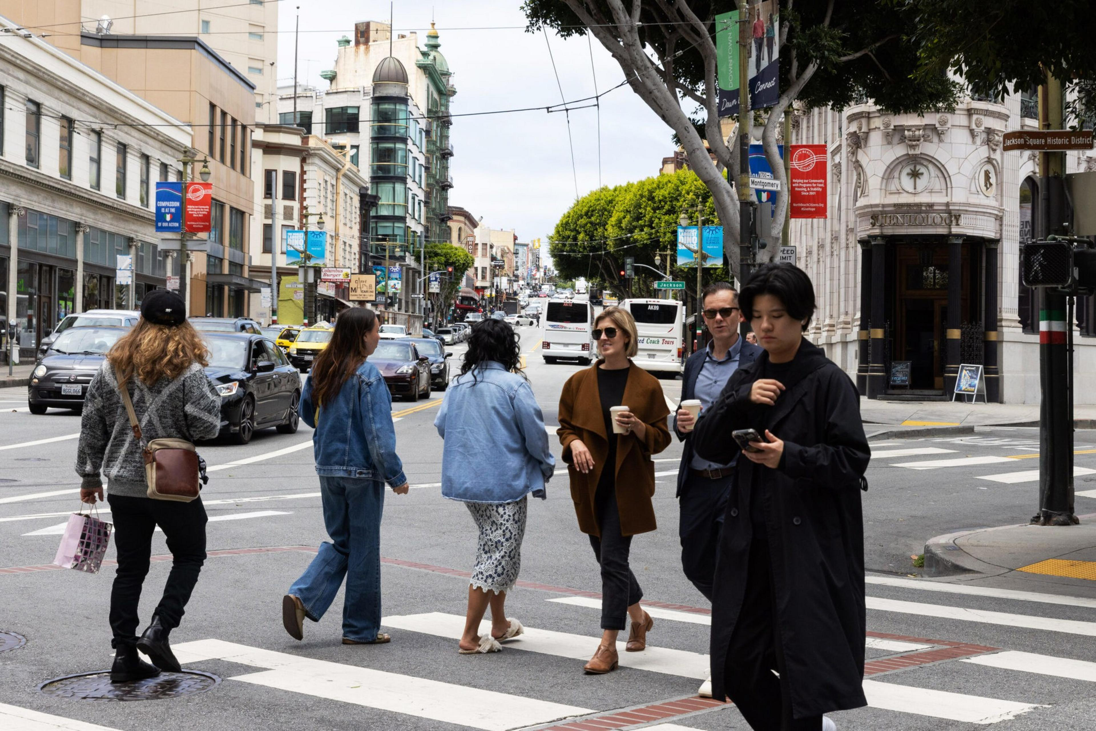 People cross a city street with cars and buildings in the background.
