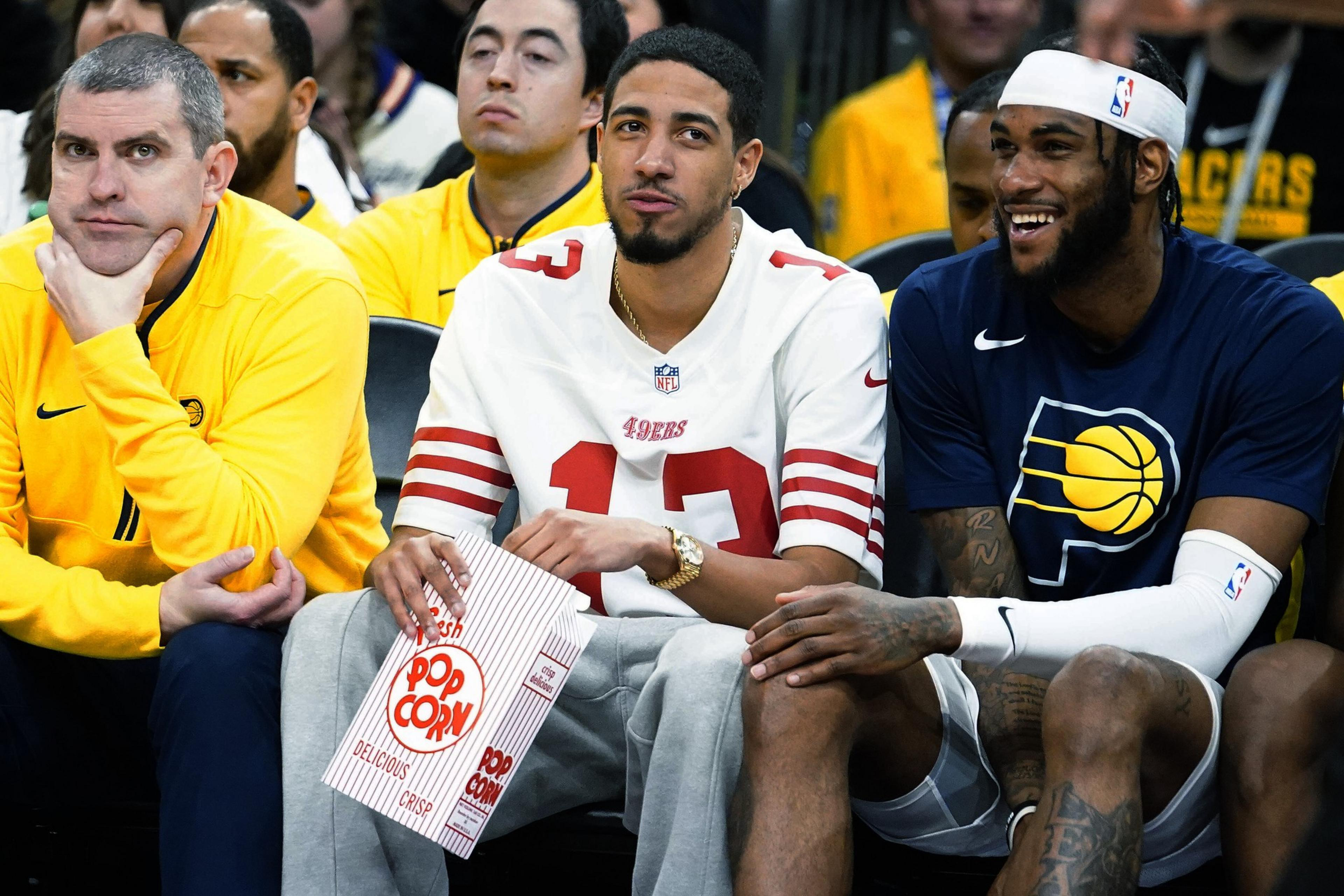 Two men are seated at a sports event, one in a 49ers jersey holding popcorn, the other in a Pacers shirt, smiling. People in yellow shirts surround them.