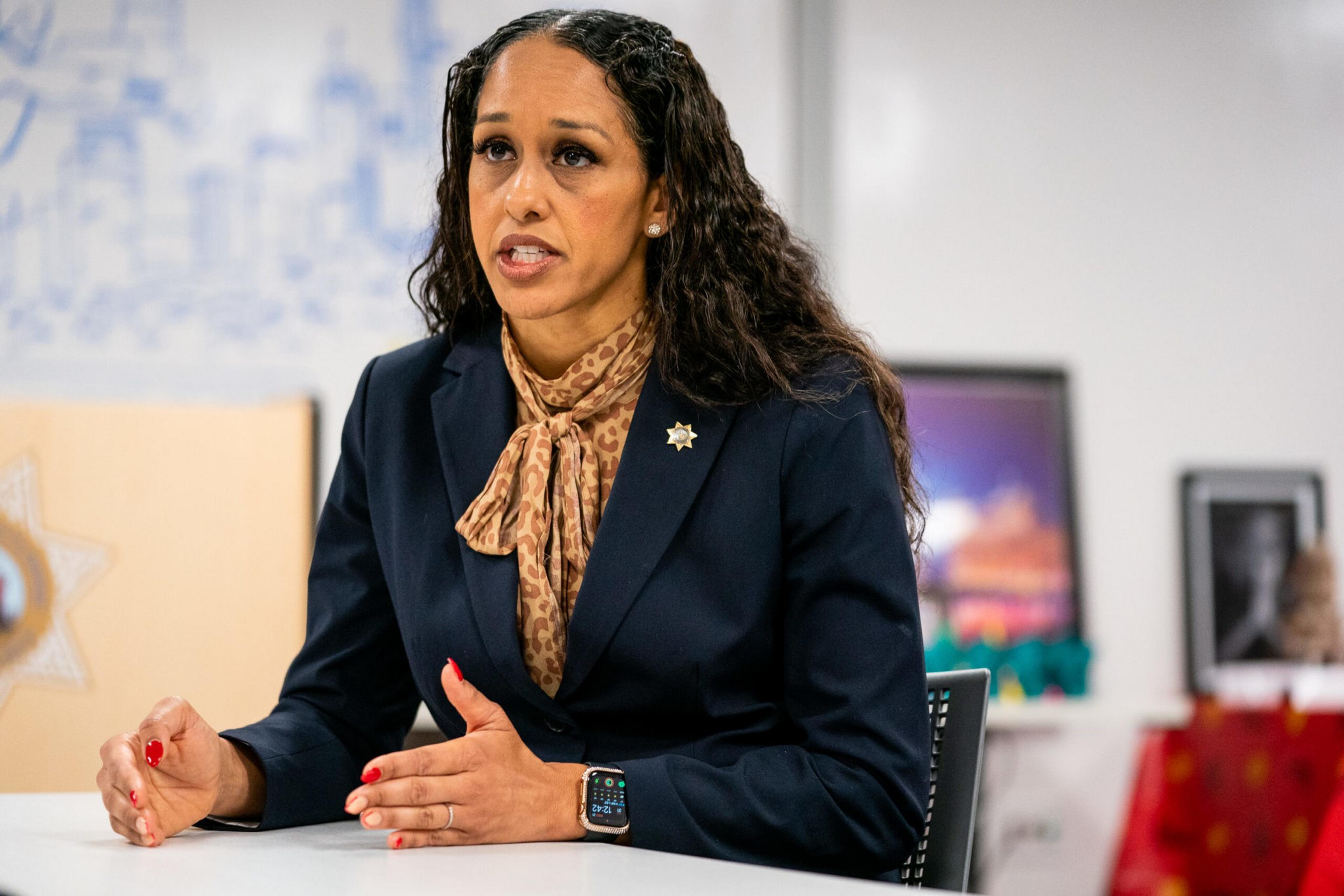 A woman in a navy blazer and patterned scarf sits at a table, speaking, with a badge on her lapel.