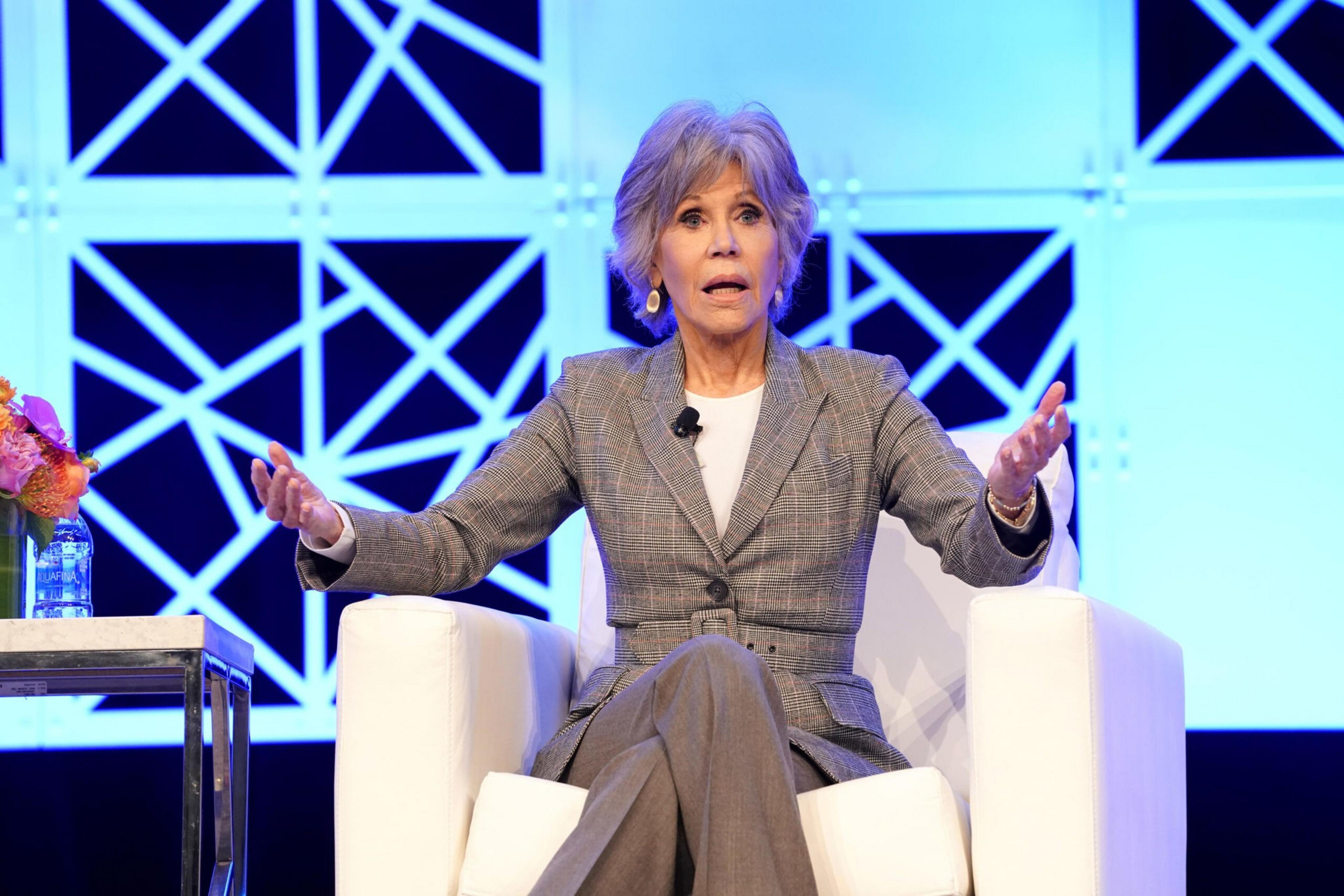 A woman with short gray hair speaks animatedly while sitting on a white chair, wearing a gray plaid suit with a white shirt, against a blue geometric backdrop.