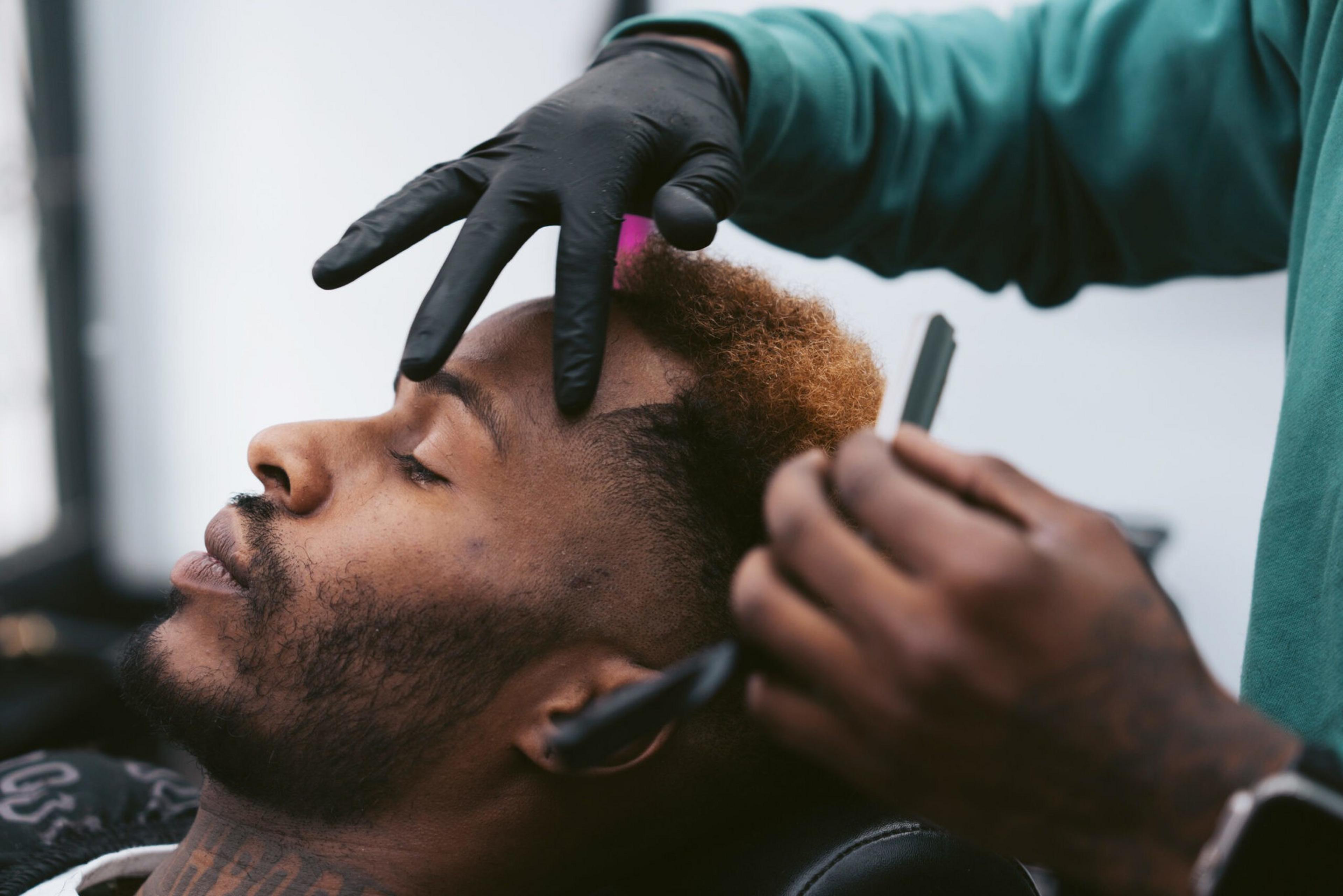 A man with closed eyes is getting a haircut while the barber wears black gloves and holds a razor near his hairline.