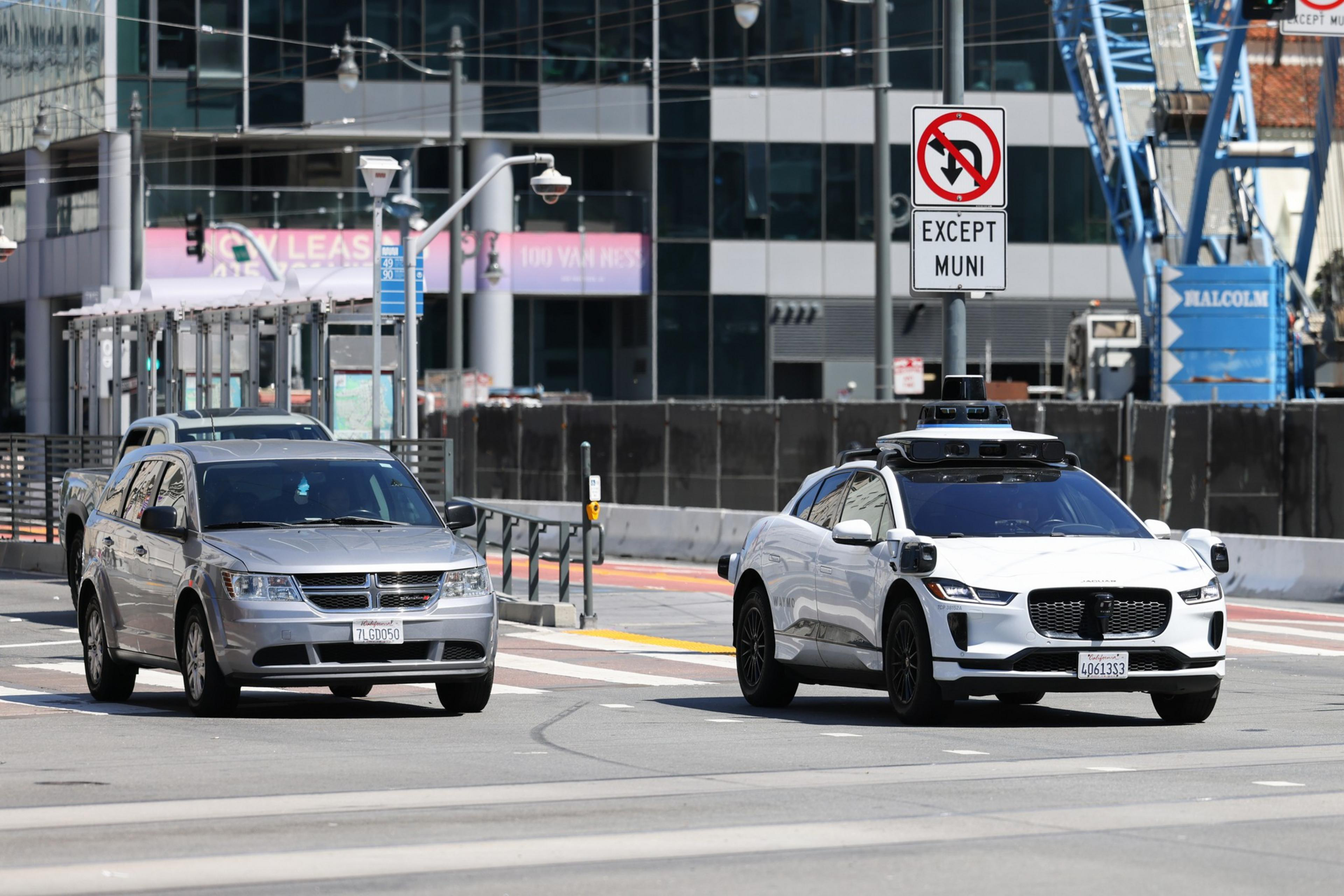 A silver SUV and a white self-driving car with sensors are on a city street near a "No Left Turn Except Muni" sign, with modern buildings in the background.