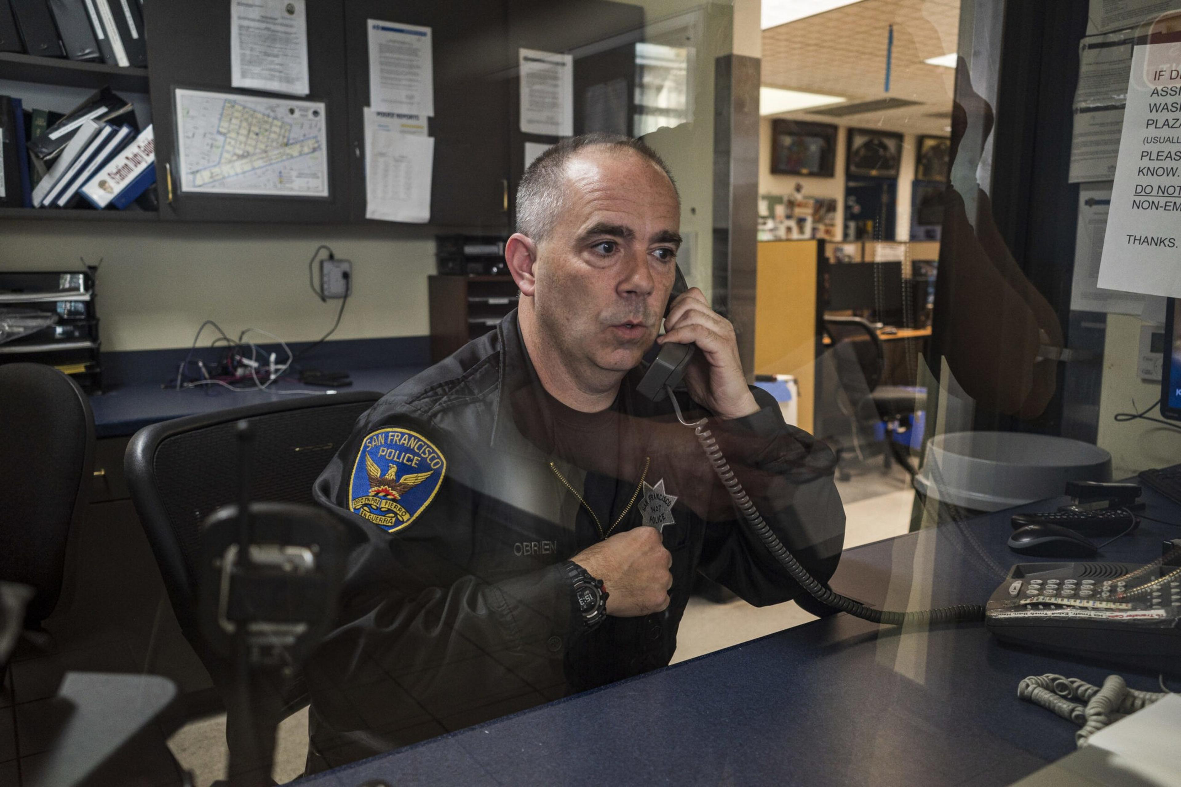 A police officer in a San Francisco Police jacket is speaking on a corded phone behind a glass partition at a desk in a police station.