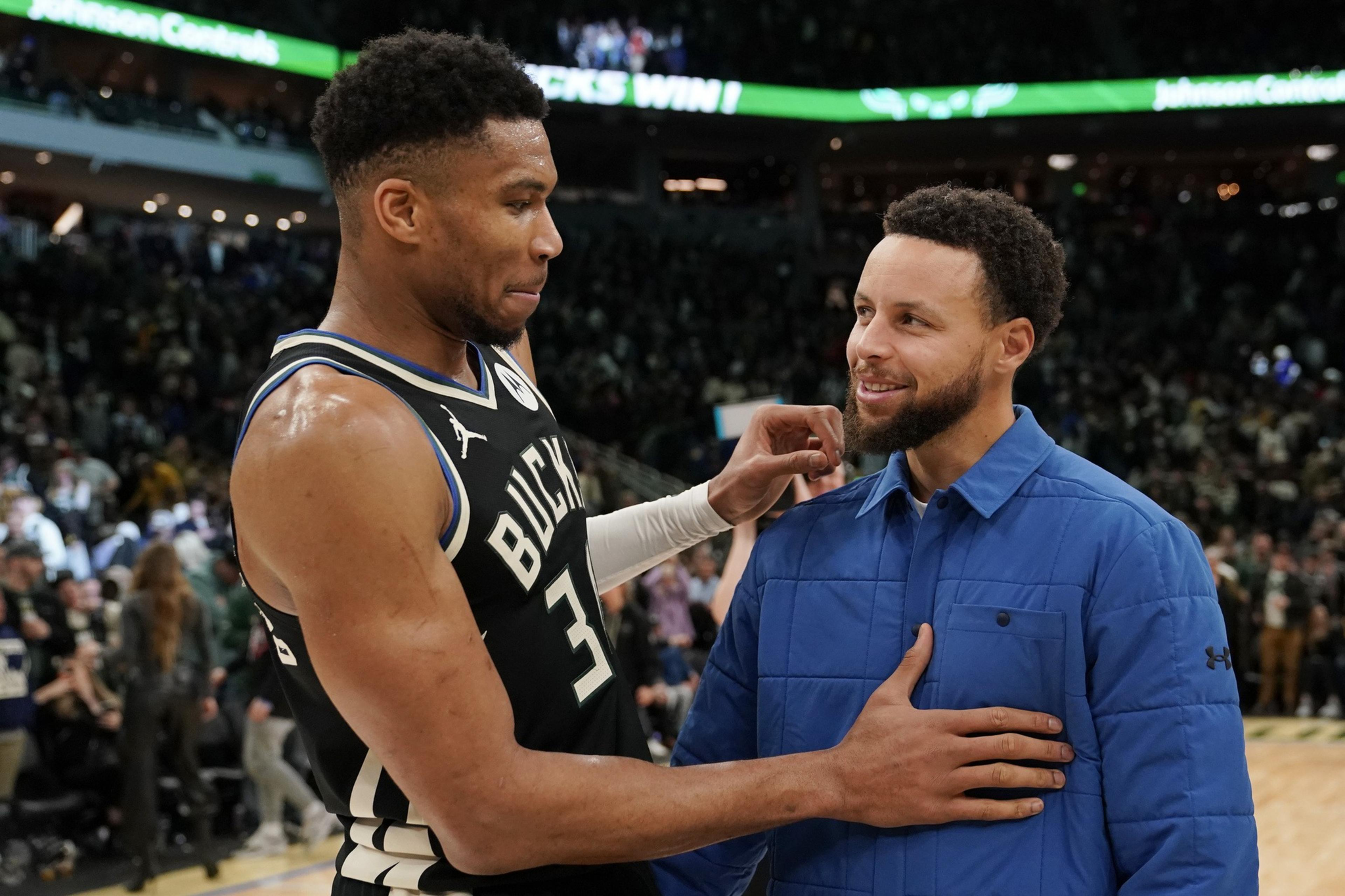 A basketball player in a Bucks jersey and a man in a blue jacket share a friendly moment on a busy indoor court.