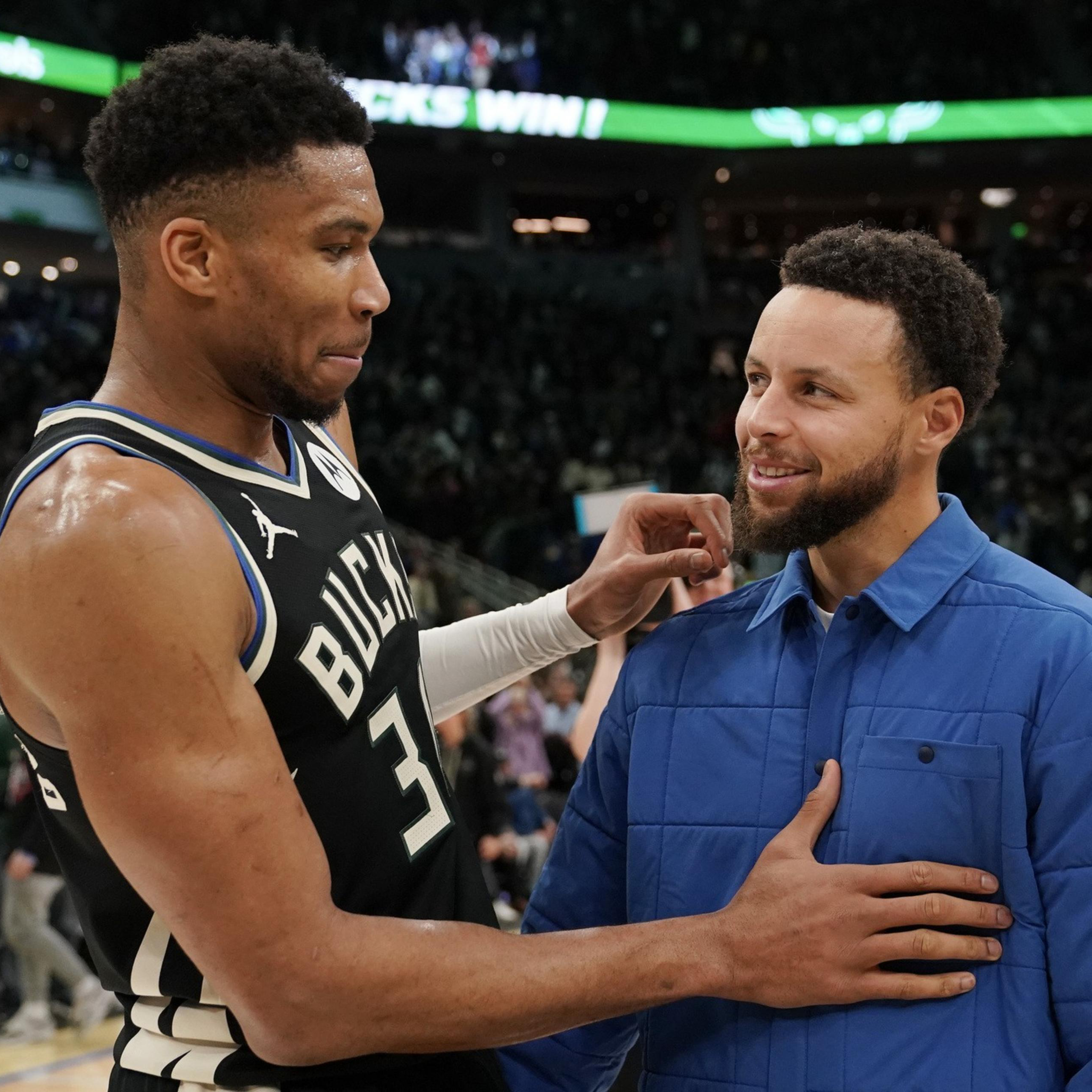 A basketball player in a Bucks jersey and a man in a blue jacket share a friendly moment on a busy indoor court.