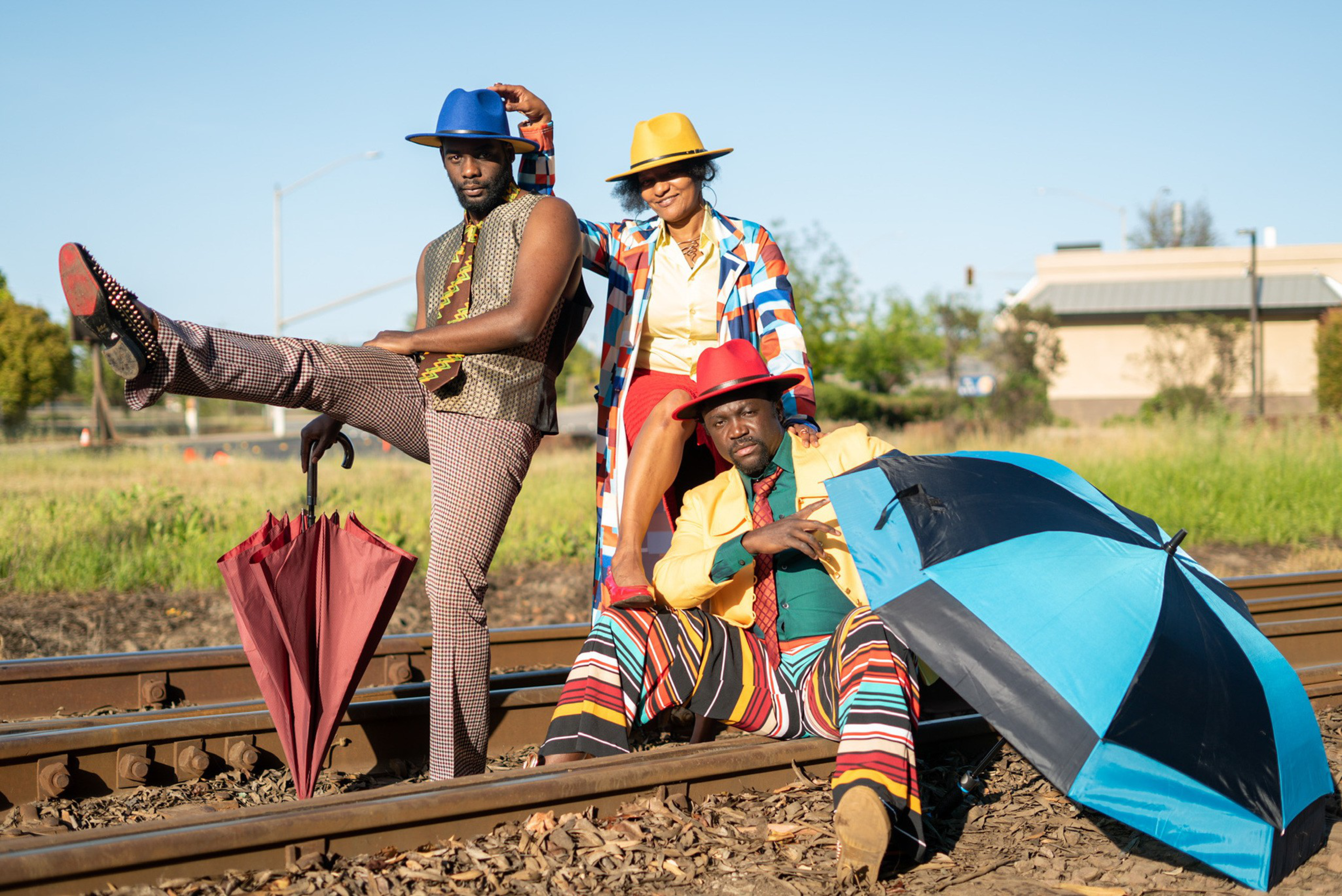 Three people in colorful, retro-style outfits and hats pose on railroad tracks, holding umbrellas, with a grassy field and buildings in the background.
