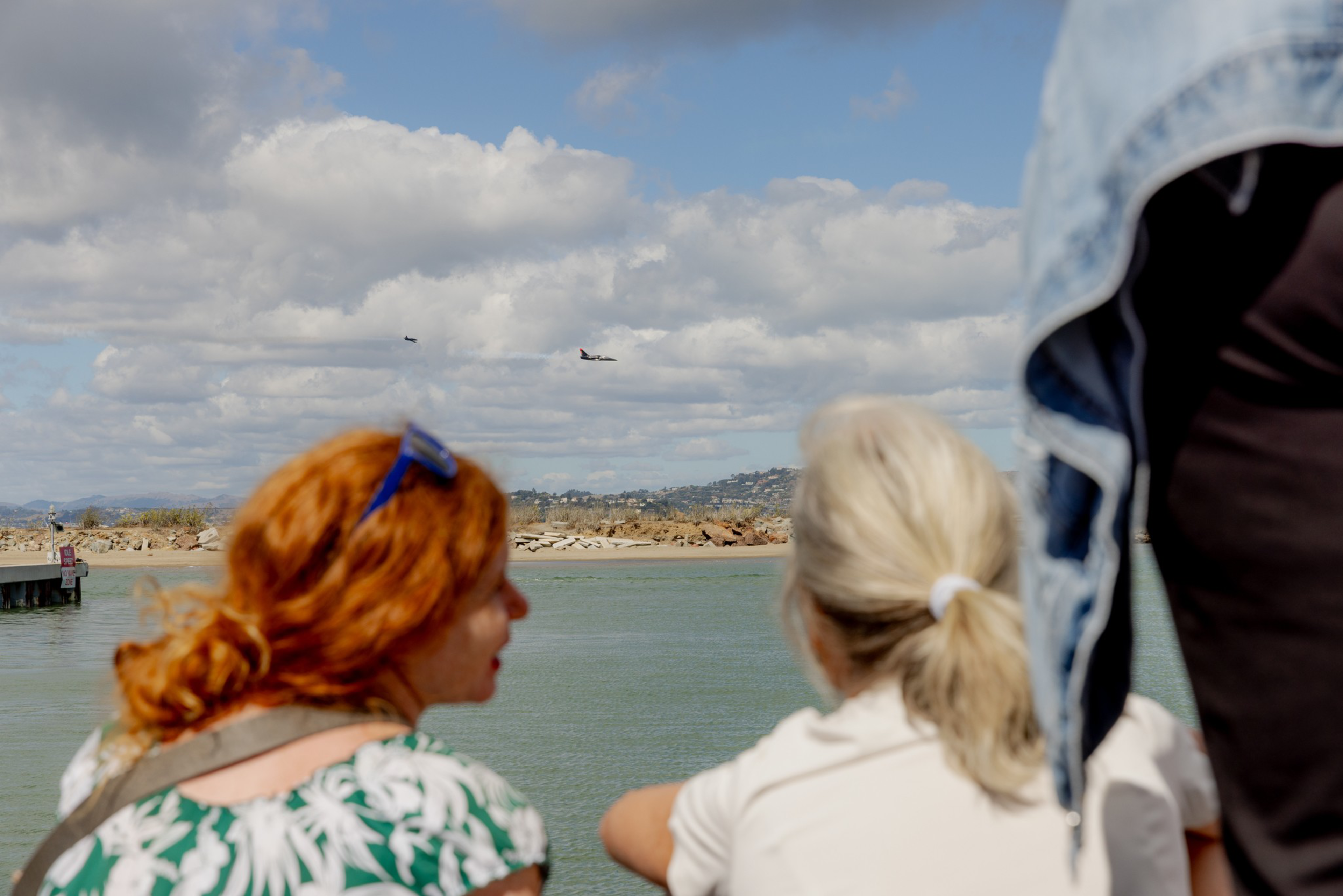 Two people with light skin, one with red hair and one with blonde, watch military planes flying over water under a partly cloudy sky.