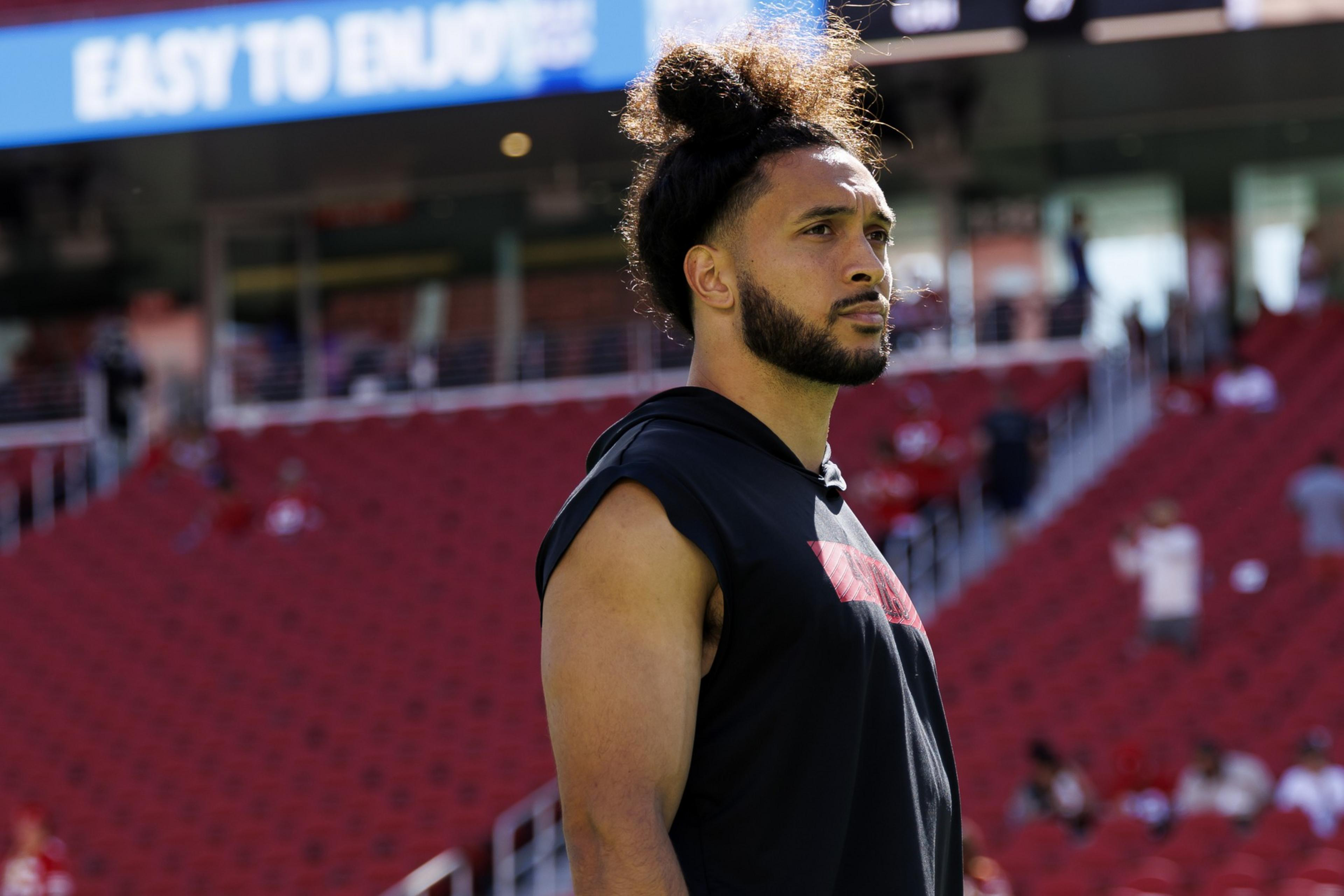 A focused athlete with tied-up curly hair and a short beard wearing a sleeveless black hoodie stands in a stadium with red seats and a sign behind him that reads "EASY TO ENJOY."