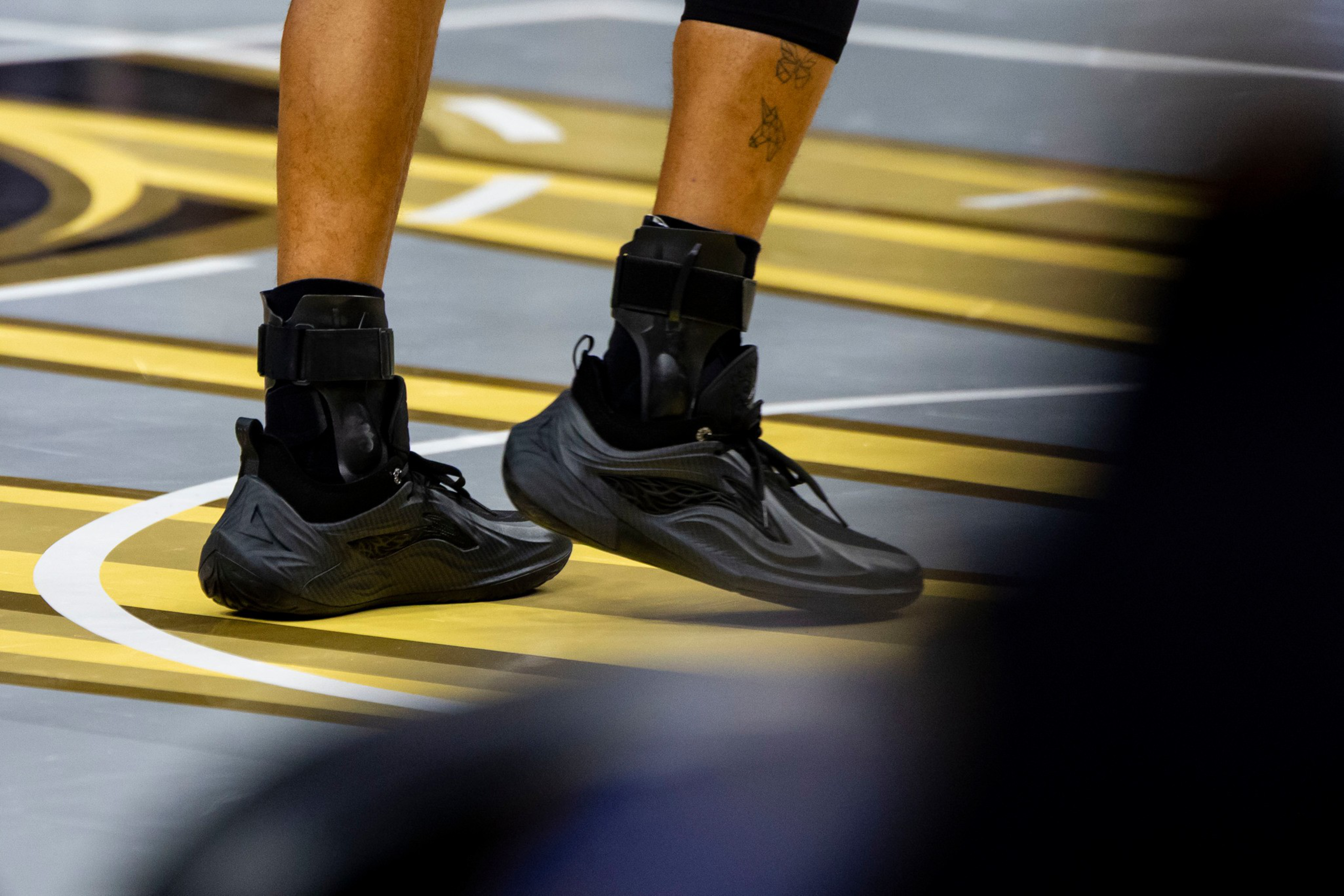 A person wearing black athletic shoes and ankle braces stands on a basketball court with gray and yellow markings.
