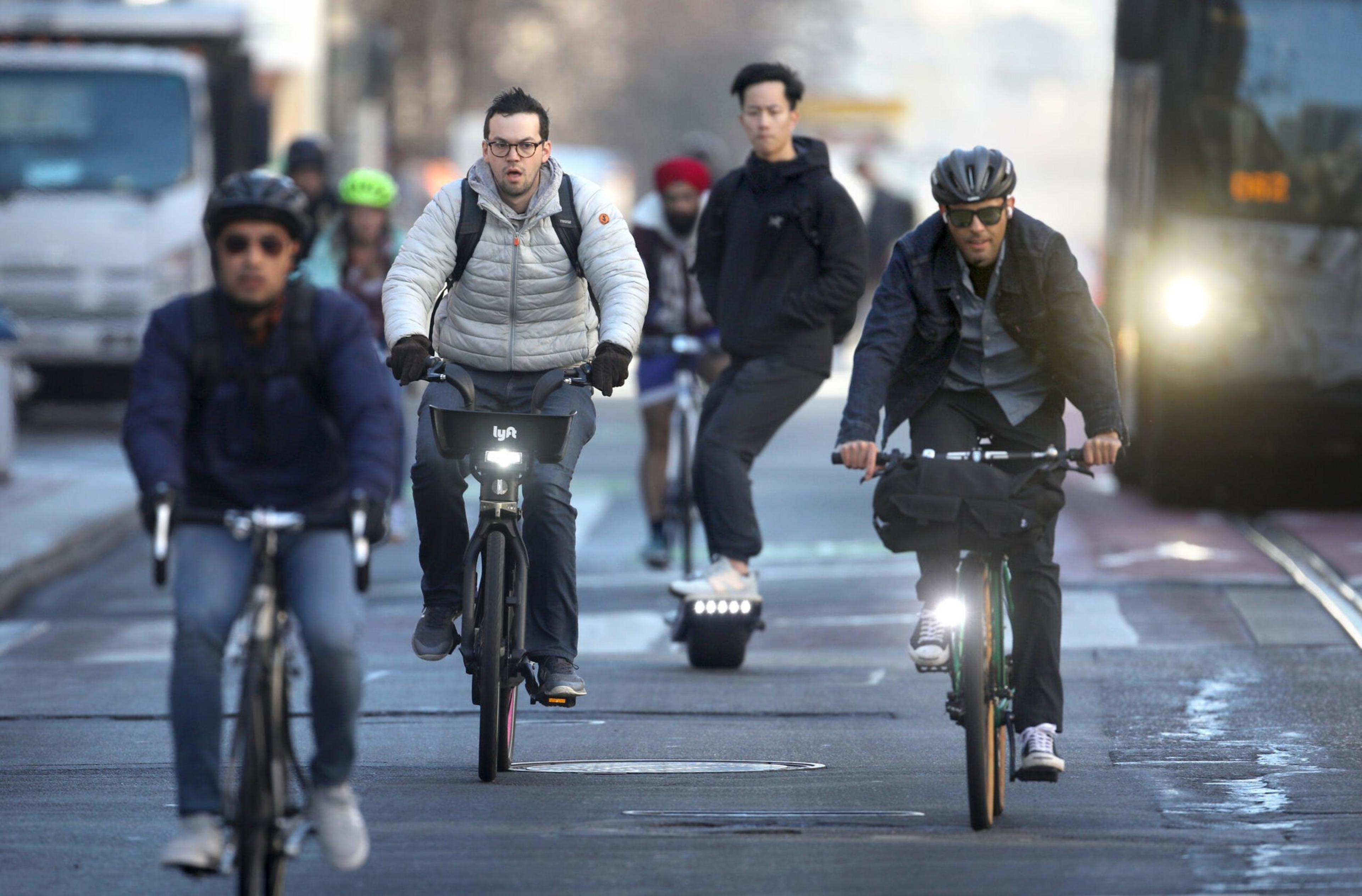 Three cyclists wearing helmets and winter clothing ride on a city street, while a pedestrian on an electric unicycle follows behind them.