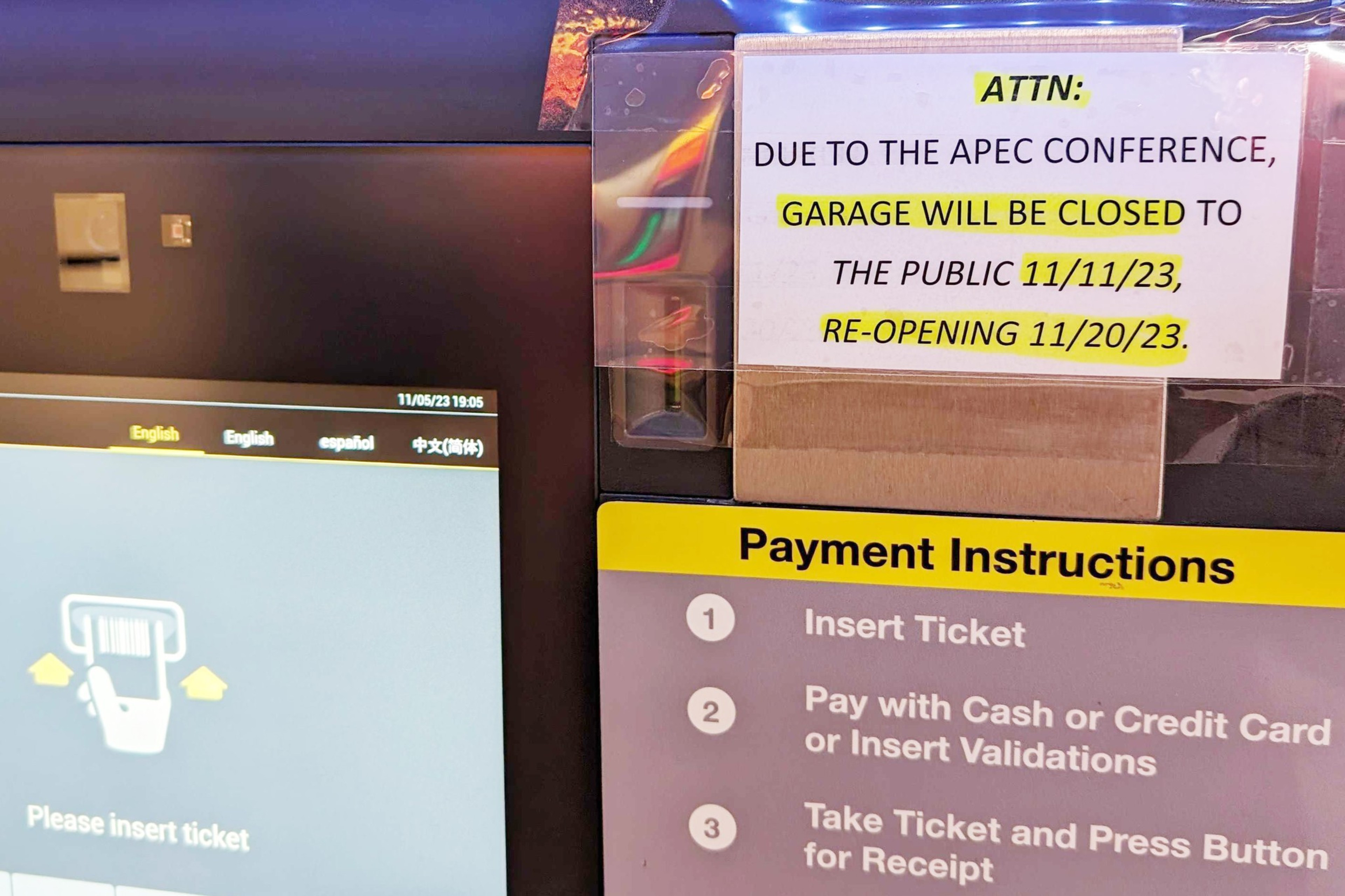 A sign is taped to a parking garage payment machine that reads &quot;ATTN: DUE TO THE APEC CONFERENCE, GARAGE WILL BE CLOSED TO THE PUBLIC 11/11/22, RE-OPENING 11/20/23.&quot; More text on the machine reads &quot;Payment Instructions&quot;, &quot;Insert Ticket&quot;, &quot;Please insert ticket&quot; among other general phrases for paying for parking.