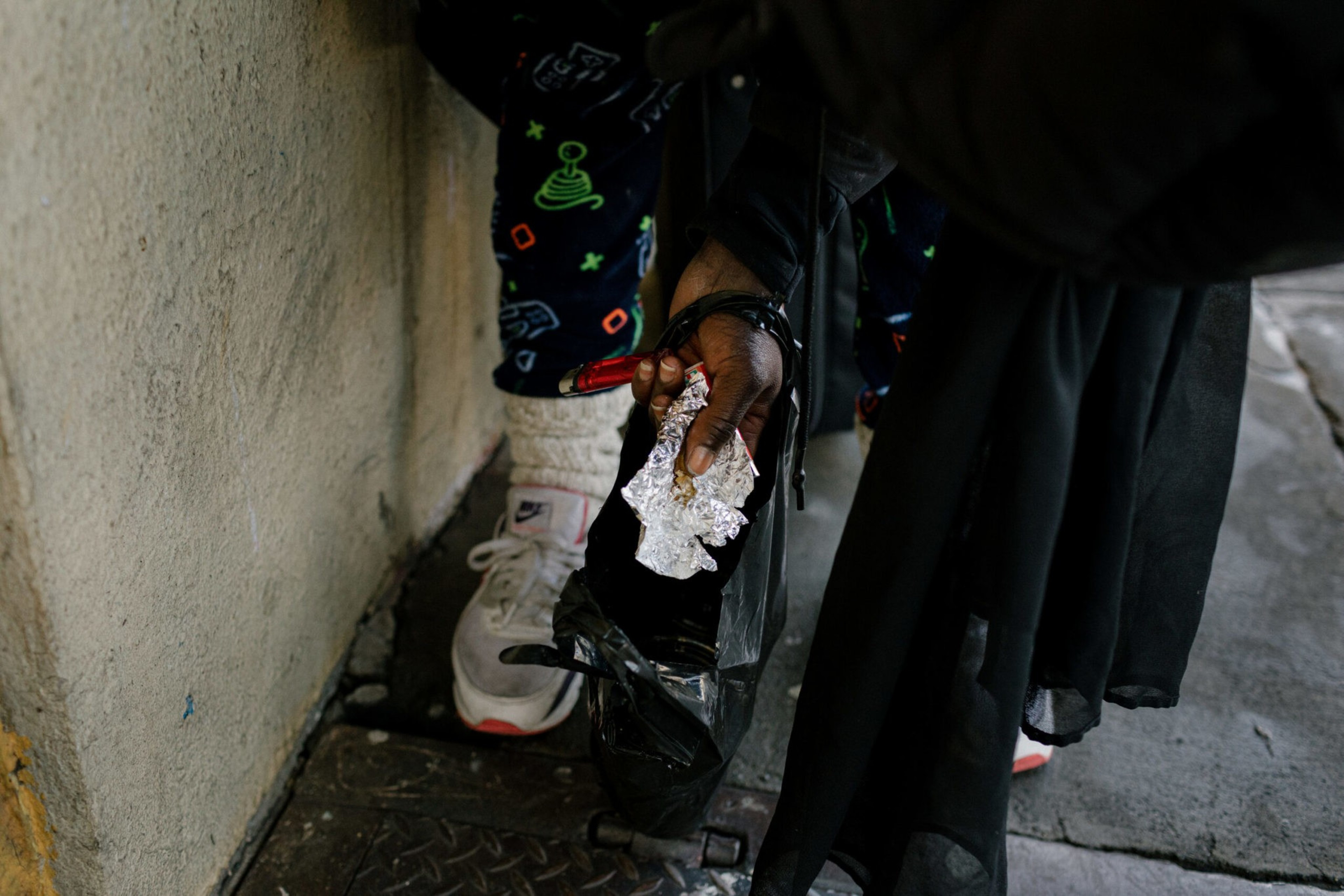 A person wearing white sneakers and dark pants with colorful designs holds a crumpled foil piece while standing near a rough wall.