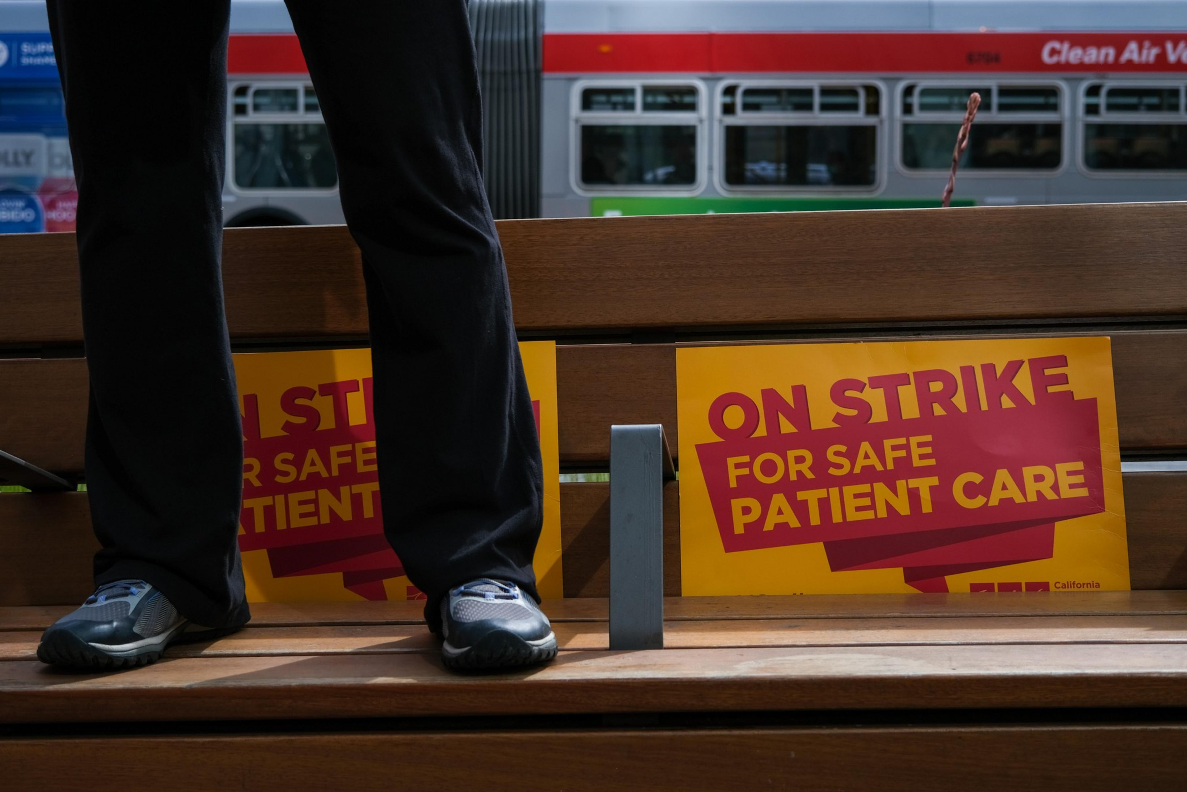 A person stands on a bench next to two yellow and red signs that read "ON STRIKE FOR SAFE PATIENT CARE," with a bus passing in the background.