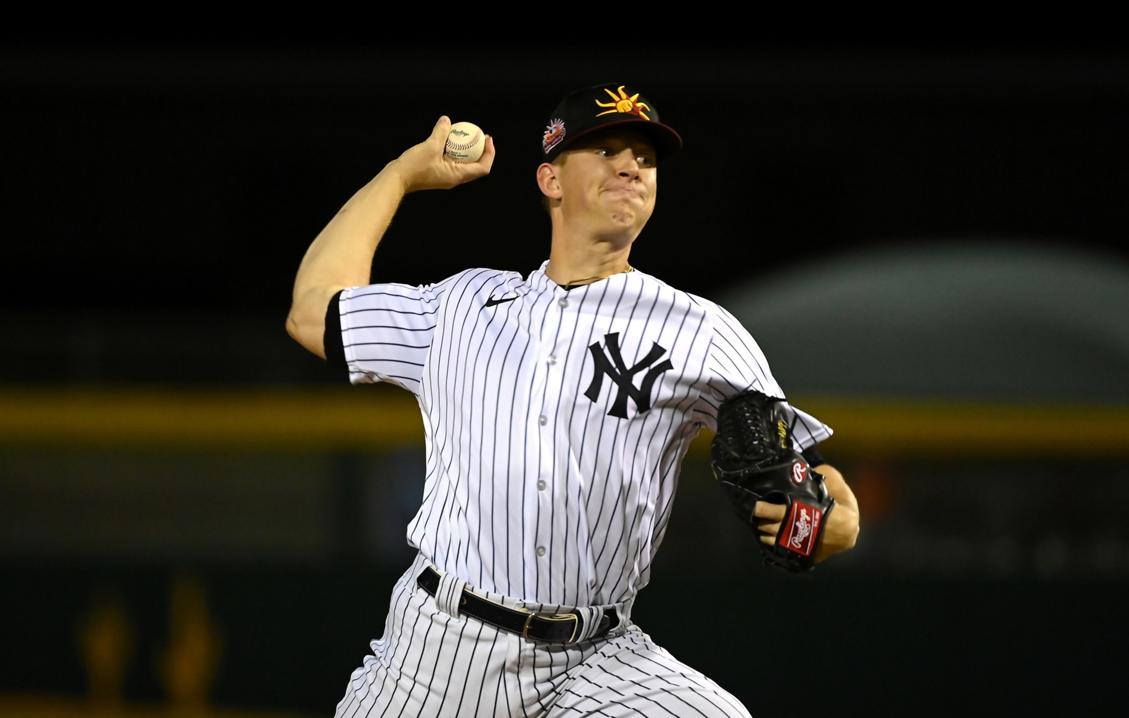 A baseball pitcher in a white pinstriped New York Yankees uniform is mid-throw, gripping a baseball with his right hand, wearing a black glove on his left.