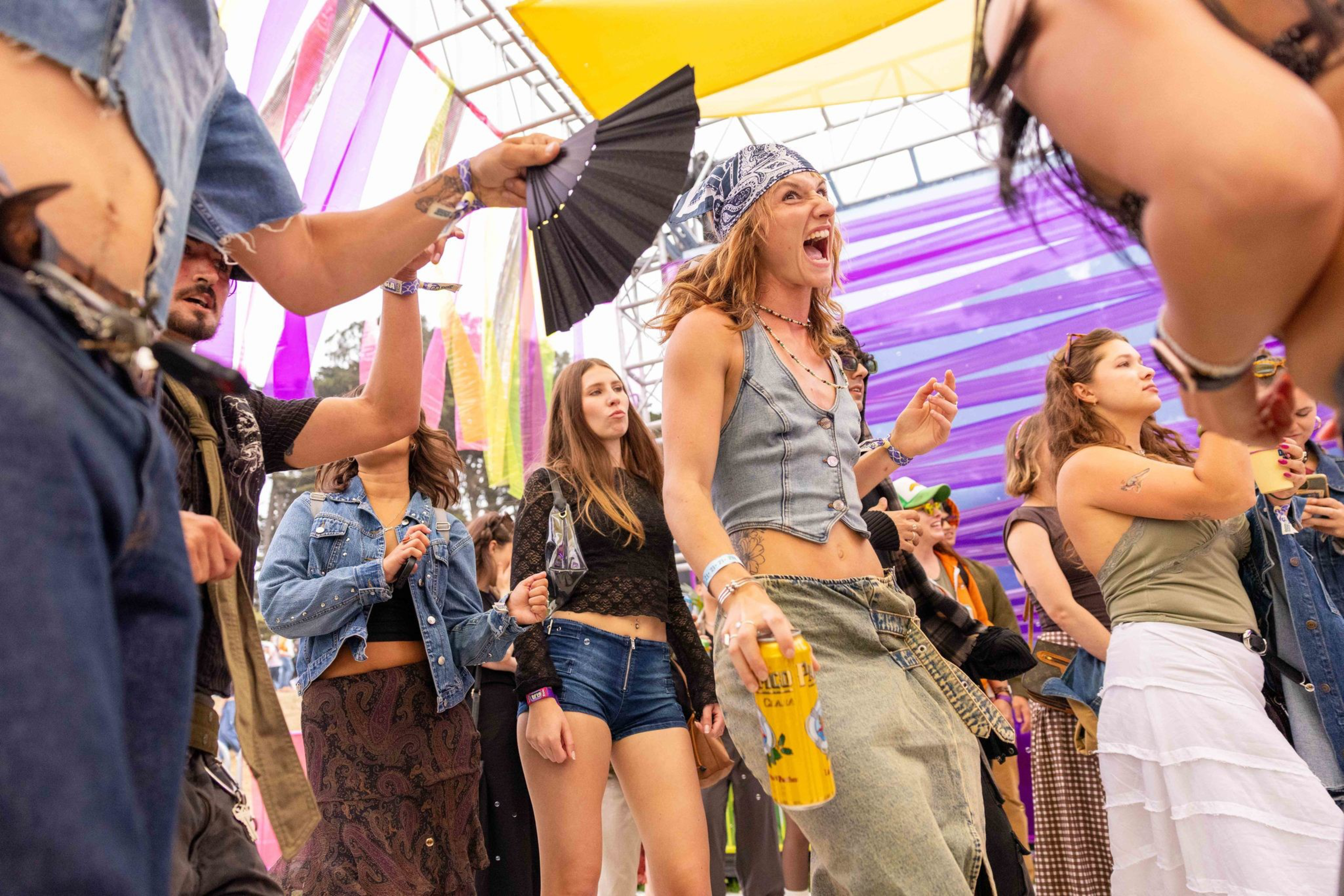 A group of diverse young people dance and smile under colorful fabric panels at an outdoor event, holding drinks and fans.