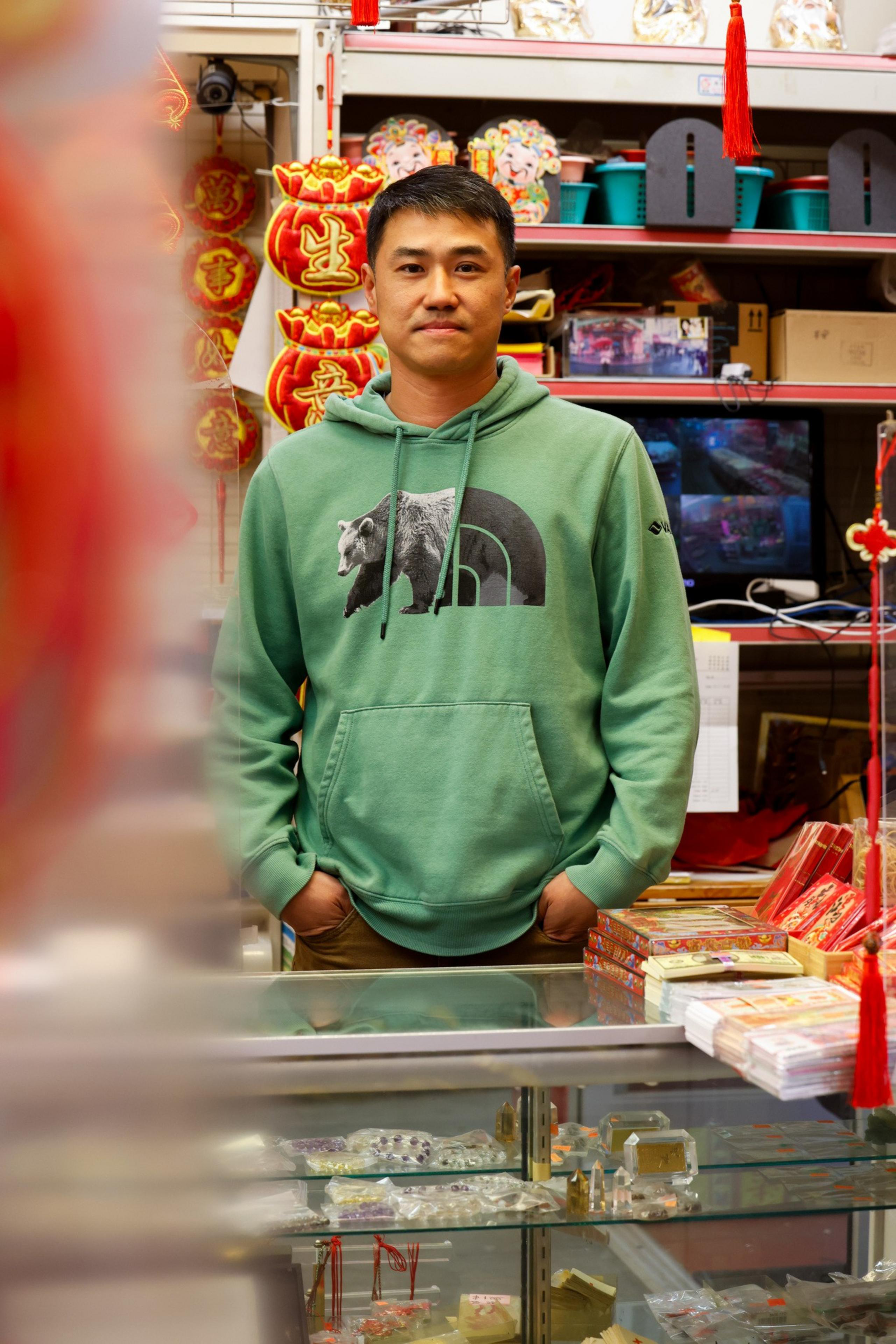 A person in a green bear hoodie stands in a shop with colorful Chinese decorations and items. Glass display cases are in the foreground.