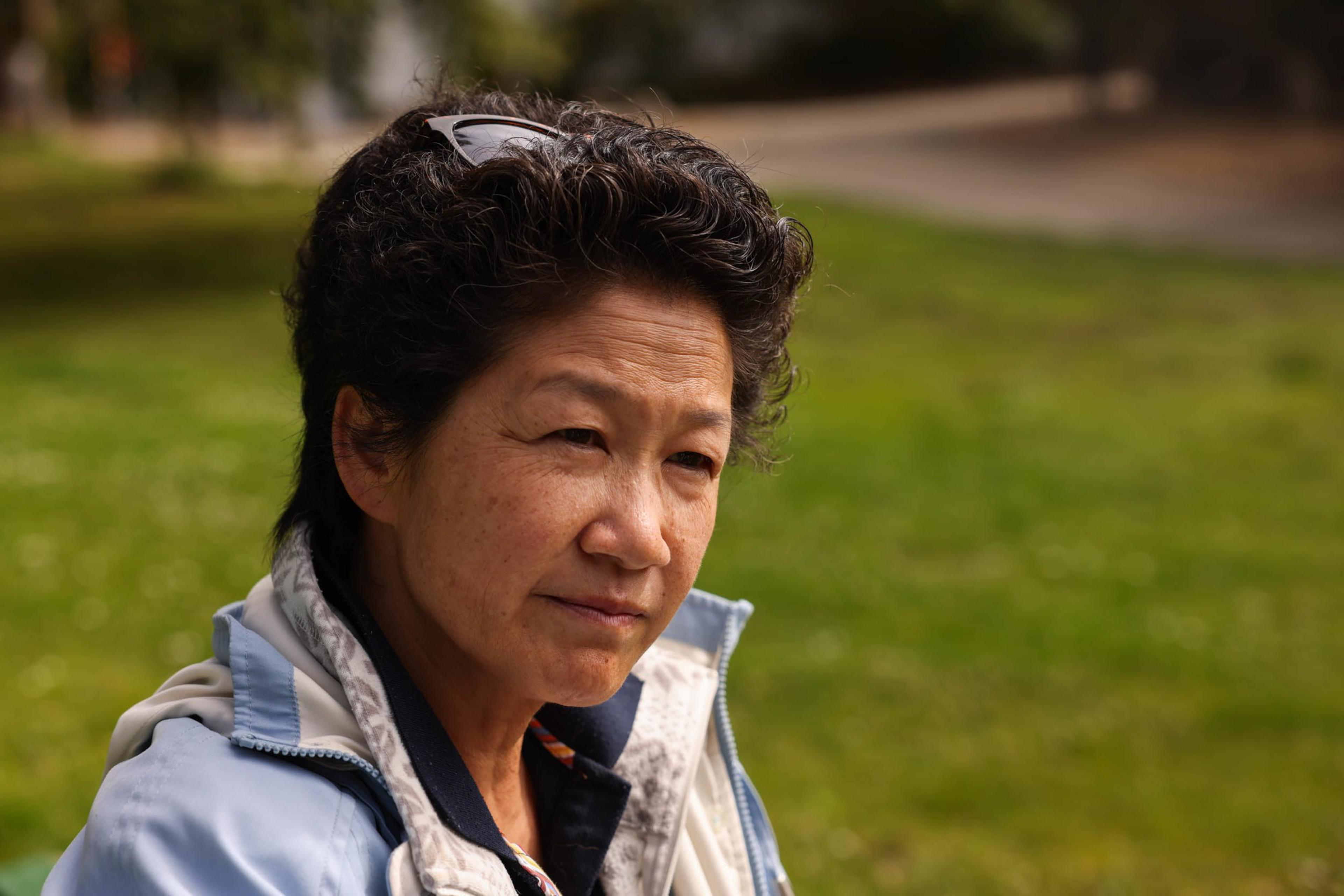 A middle-aged person with short curly hair and glasses on their head looks thoughtfully to the side, wearing a light blue jacket with a green grassy background.