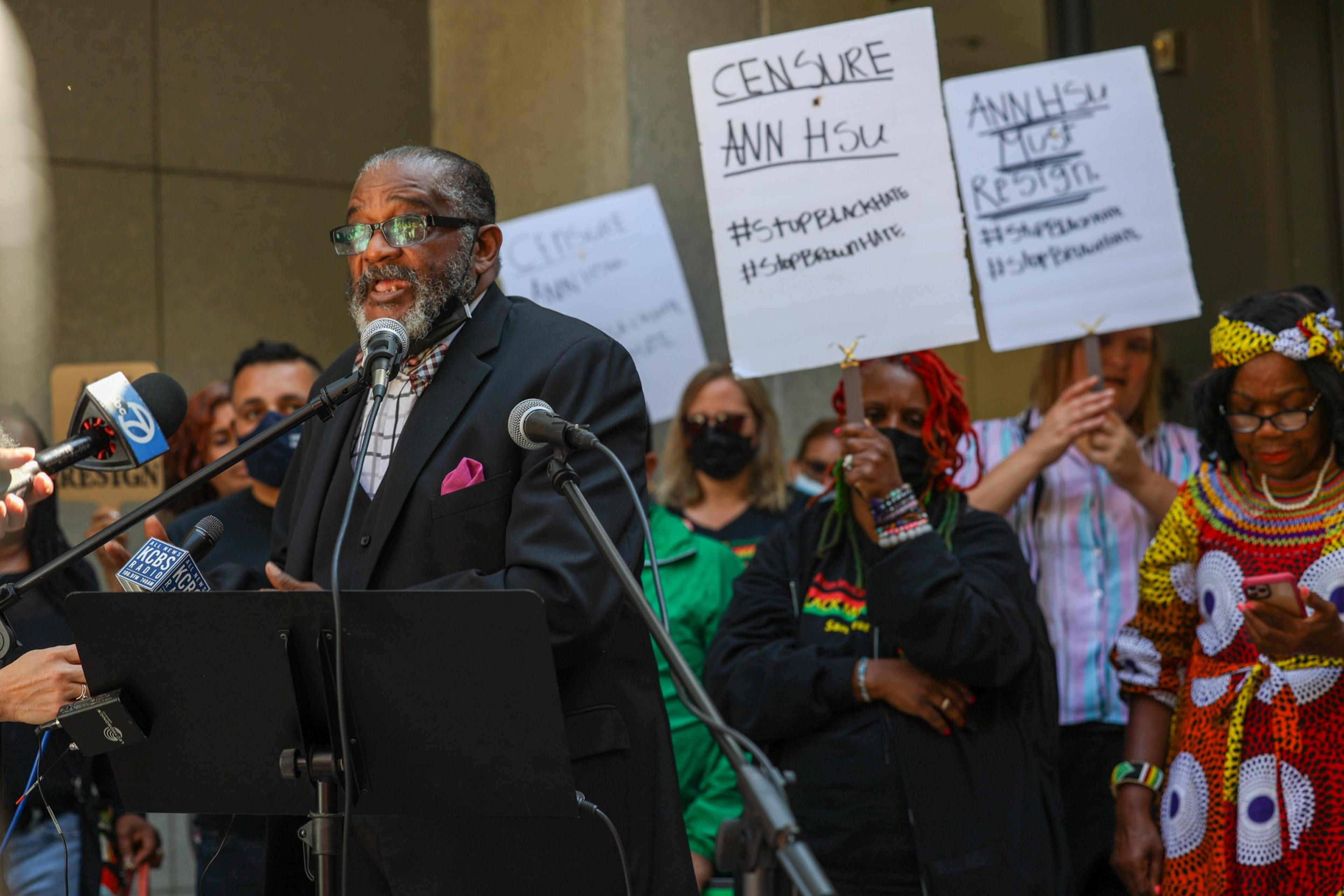 A man in a suit speaks at a microphone while people behind him hold protest signs demanding to censor Ann Hsu and stop racism.