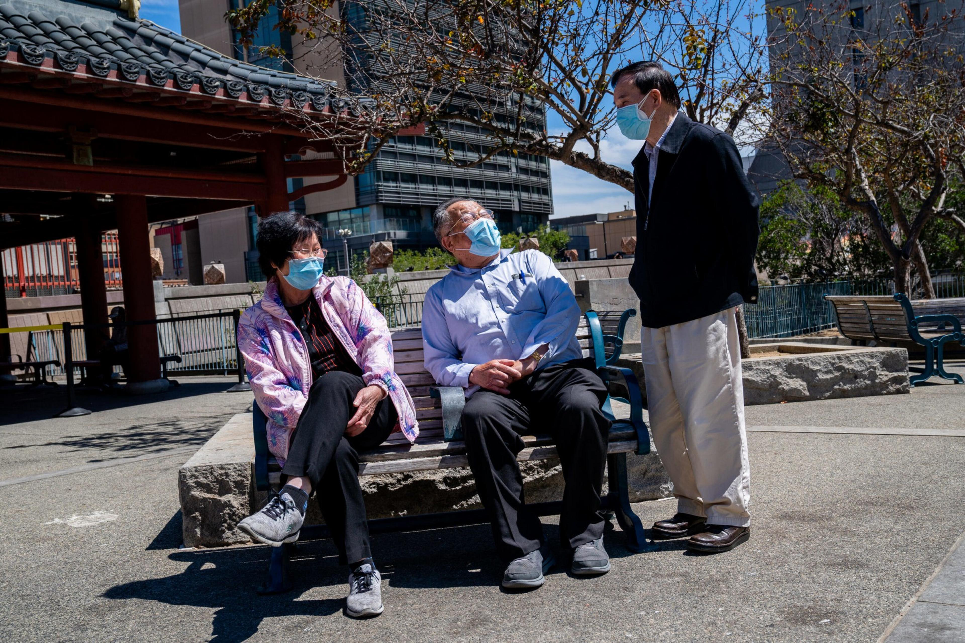 Two elderly people wearing masks sit on a bench outdoors while a third person stands nearby, also wearing a mask, in an urban park setting.