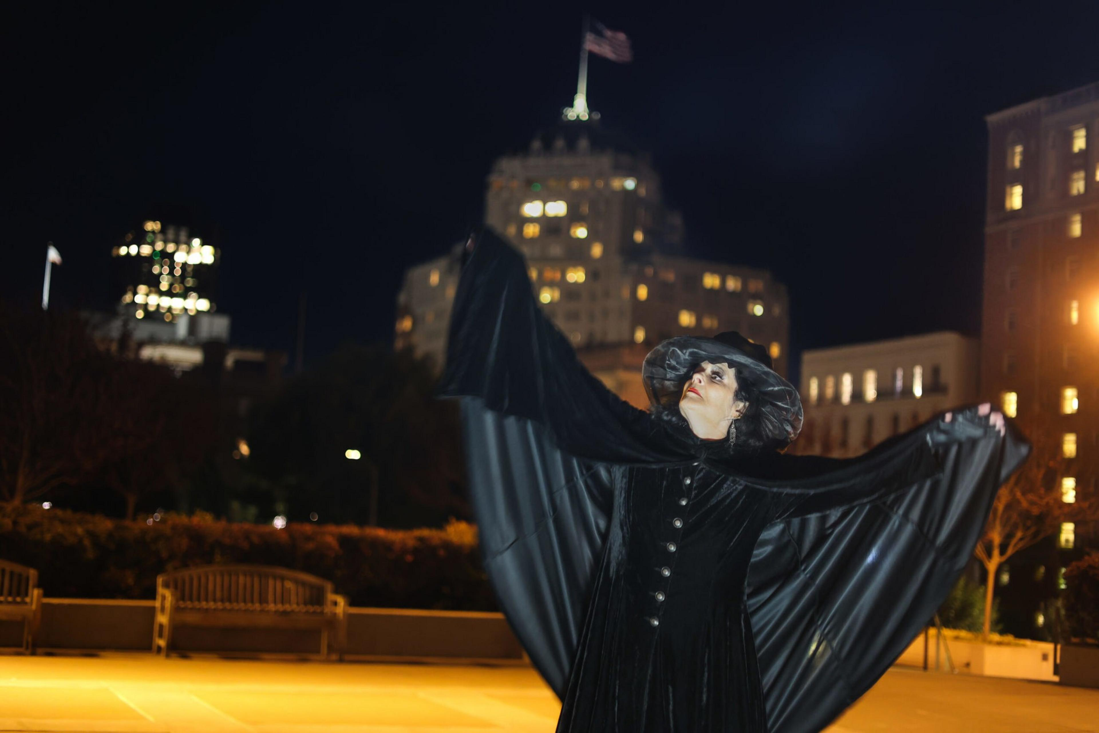 A person dressed as a witch in a black cloak and hat stretches their arms out wide in a nighttime city plaza with lit buildings in the background.