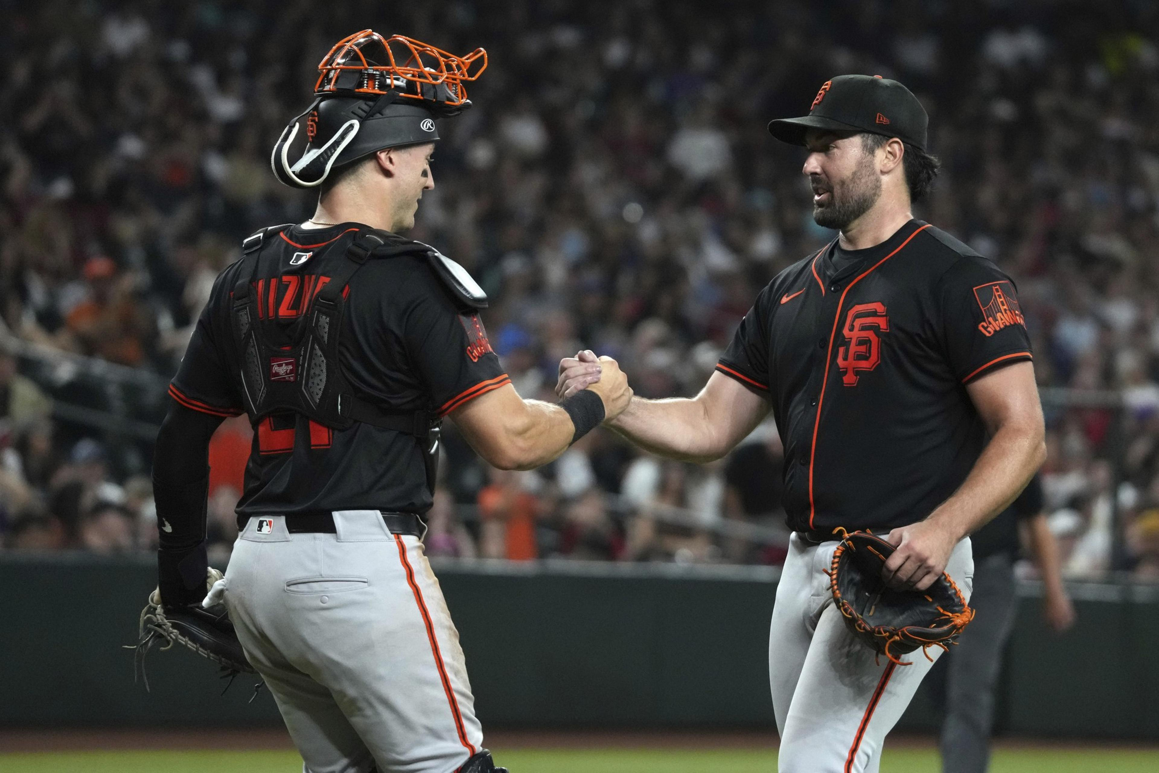 Two baseball players in black uniforms are shaking hands on the field. One wears catcher's gear, the other holds a glove. The background is filled with spectators.