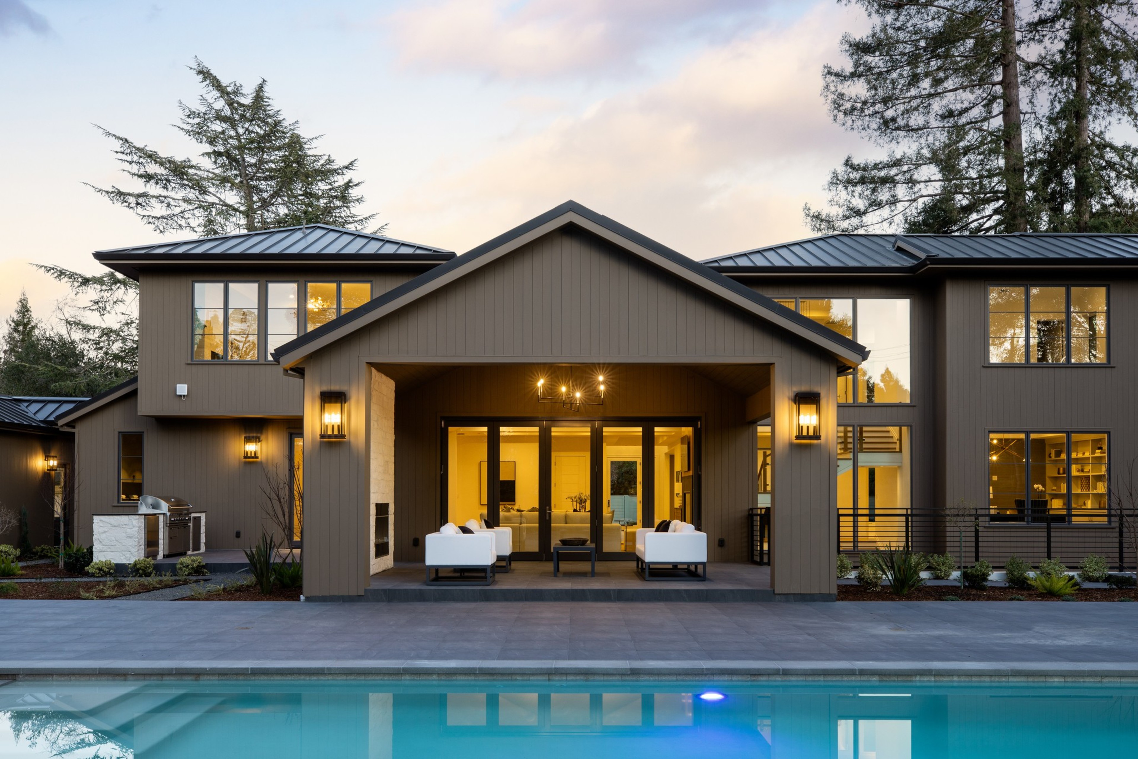 A modern two-story house with large windows, a covered patio with white sofas, and a swimming pool in the foreground during early evening.