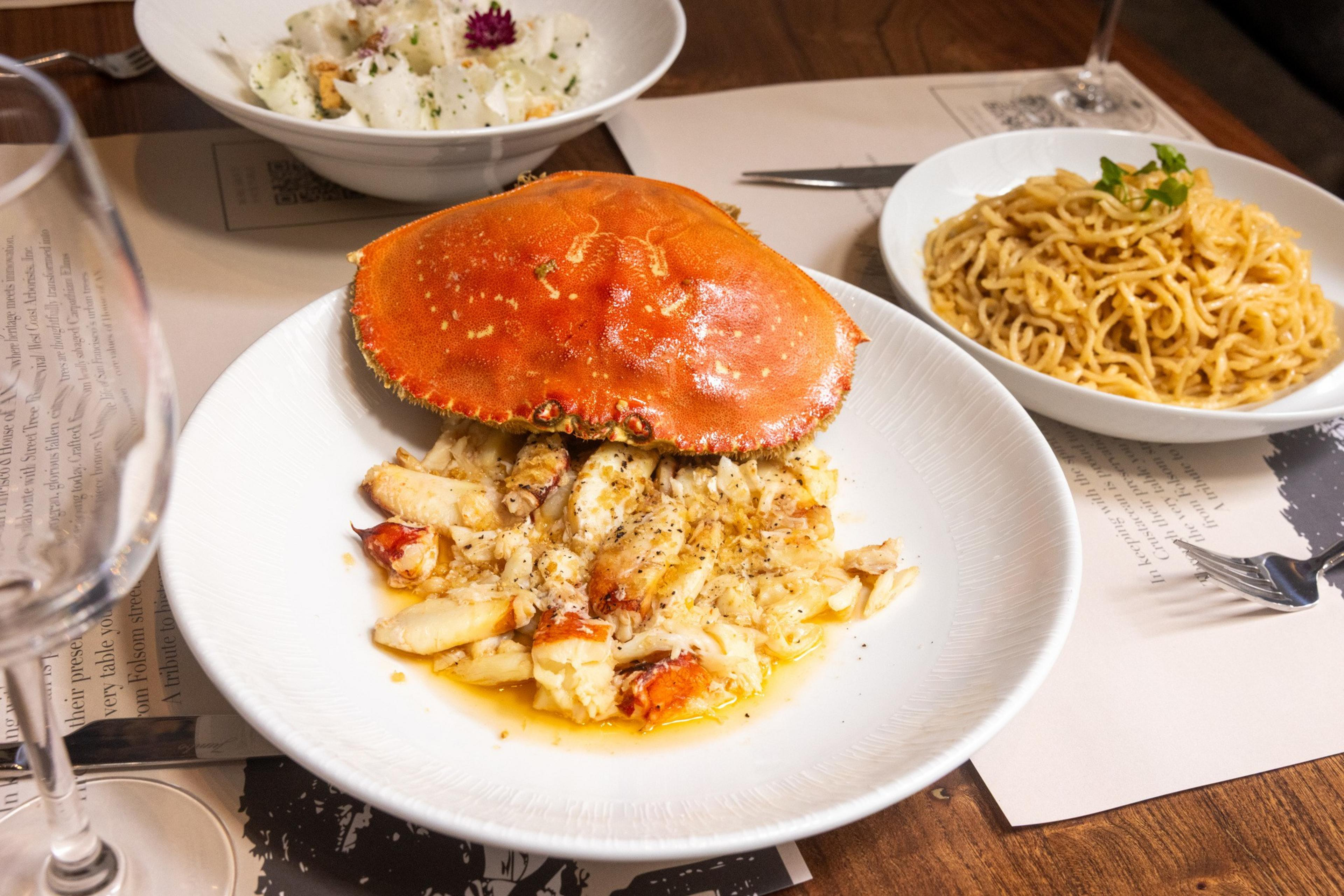 A plate of crab meat with the crab shell on top, a bowl of creamy salad, and a bowl of noodles garnished with greens on a wooden table.