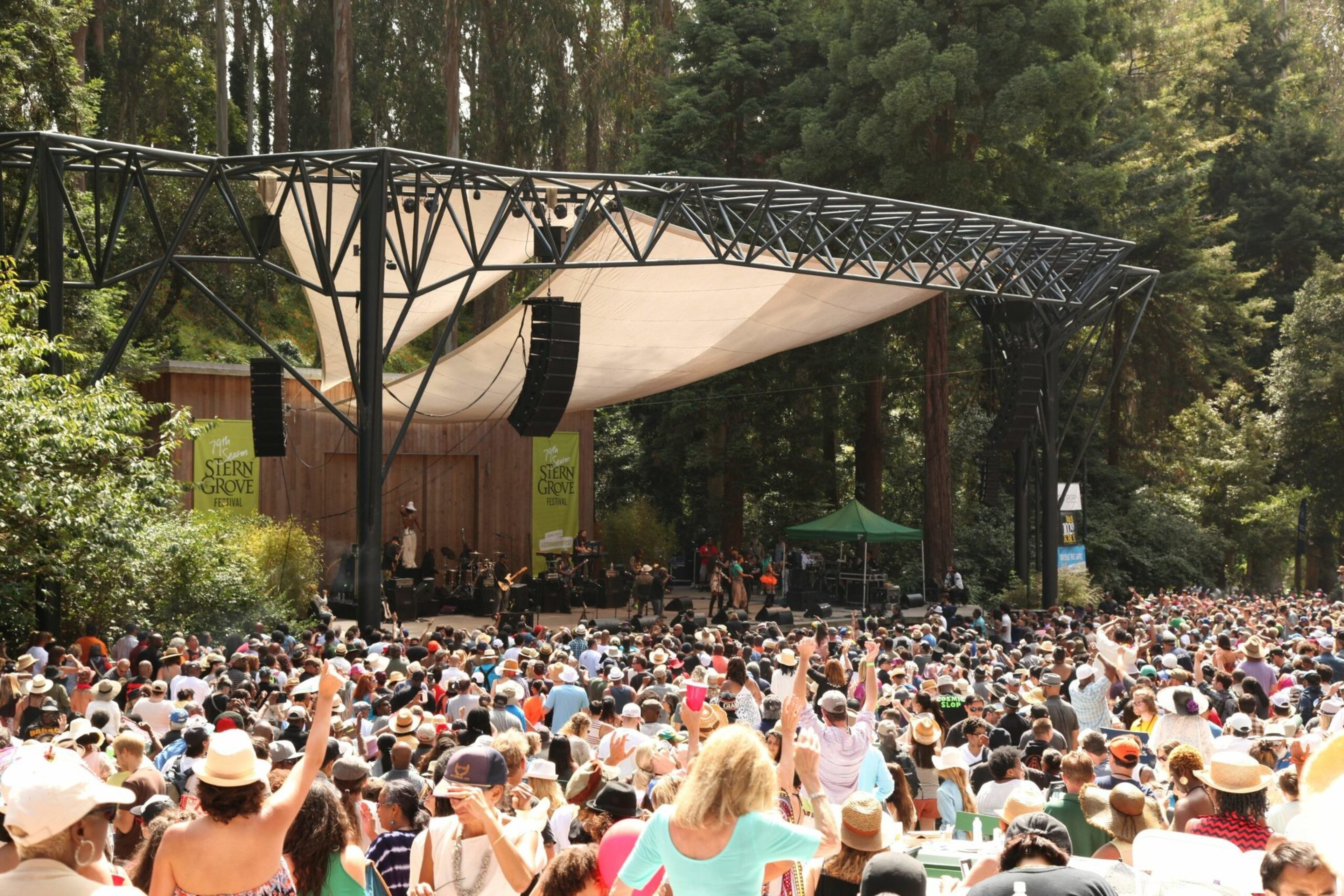 A large crowd gathers outdoors in front of a stage surrounded by tall trees, watching a live music performance at a venue called Stern Grove.