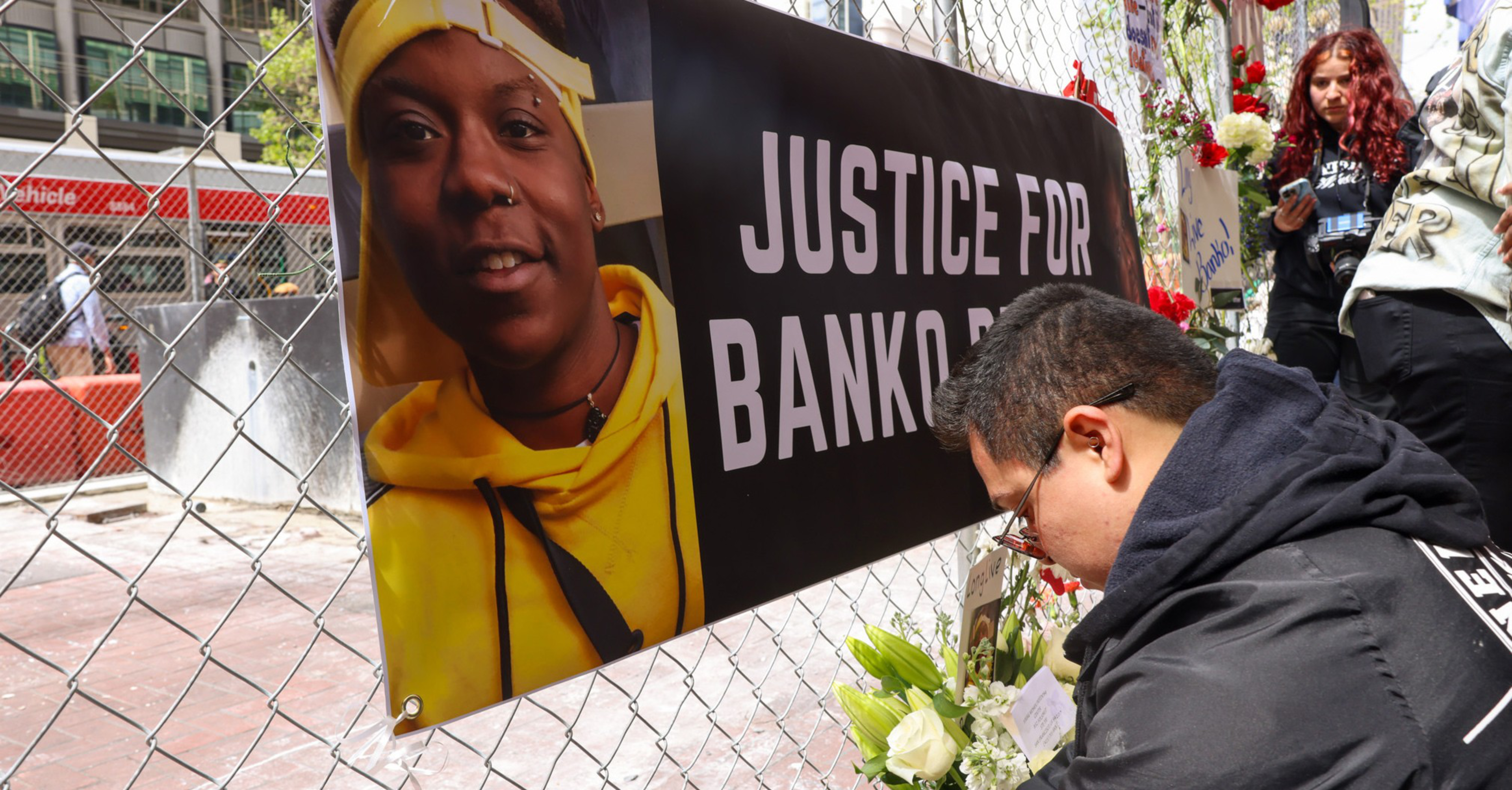 A man places flowers at a memorial next to a banner reading "Justice for Banko Rose" with her photo on a chain-link fence.