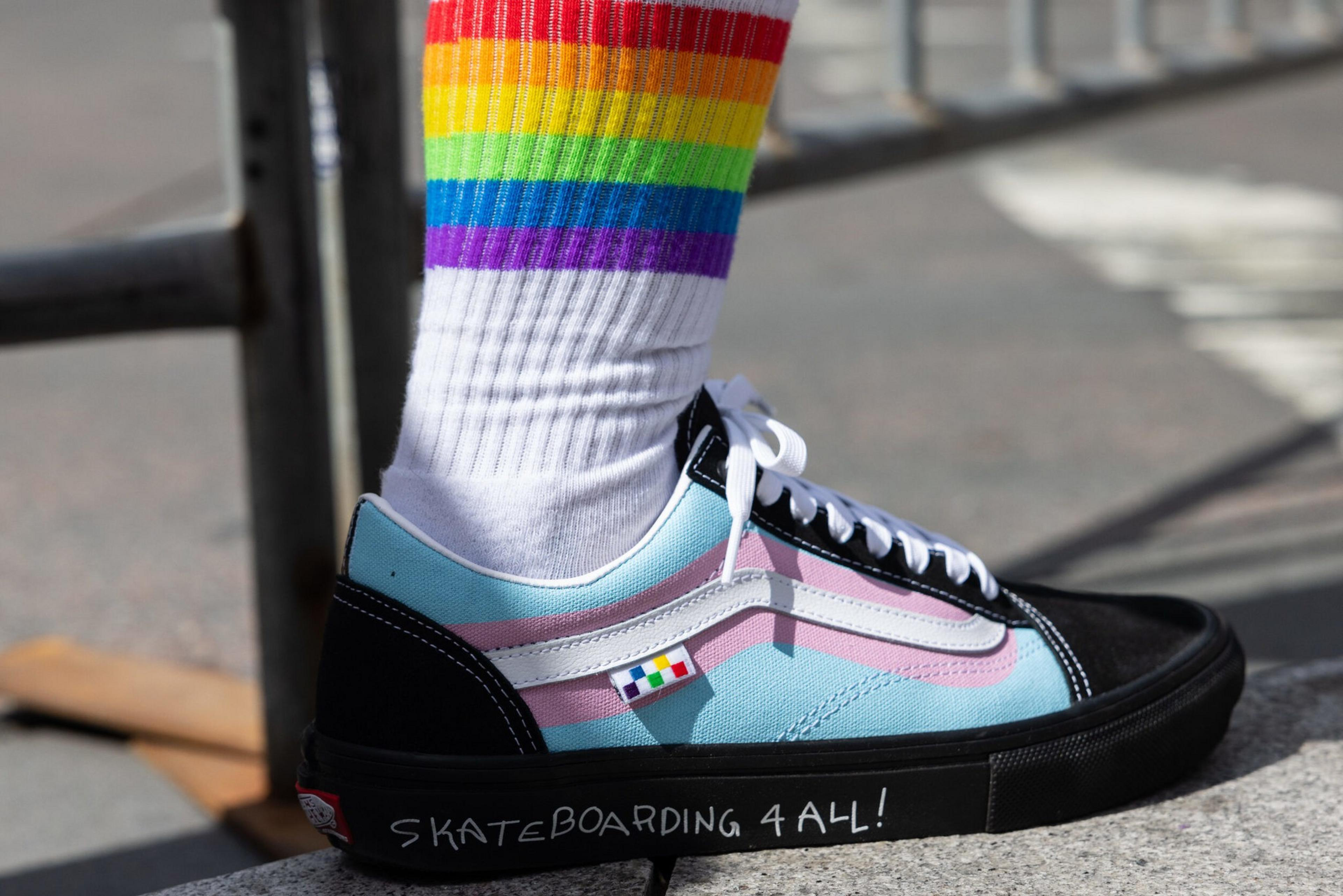 A foot wears a light blue, pink, and black skate shoe with "SKATEBOARDING 4 ALL!" written on the black sole, paired with white socks featuring rainbow stripes.