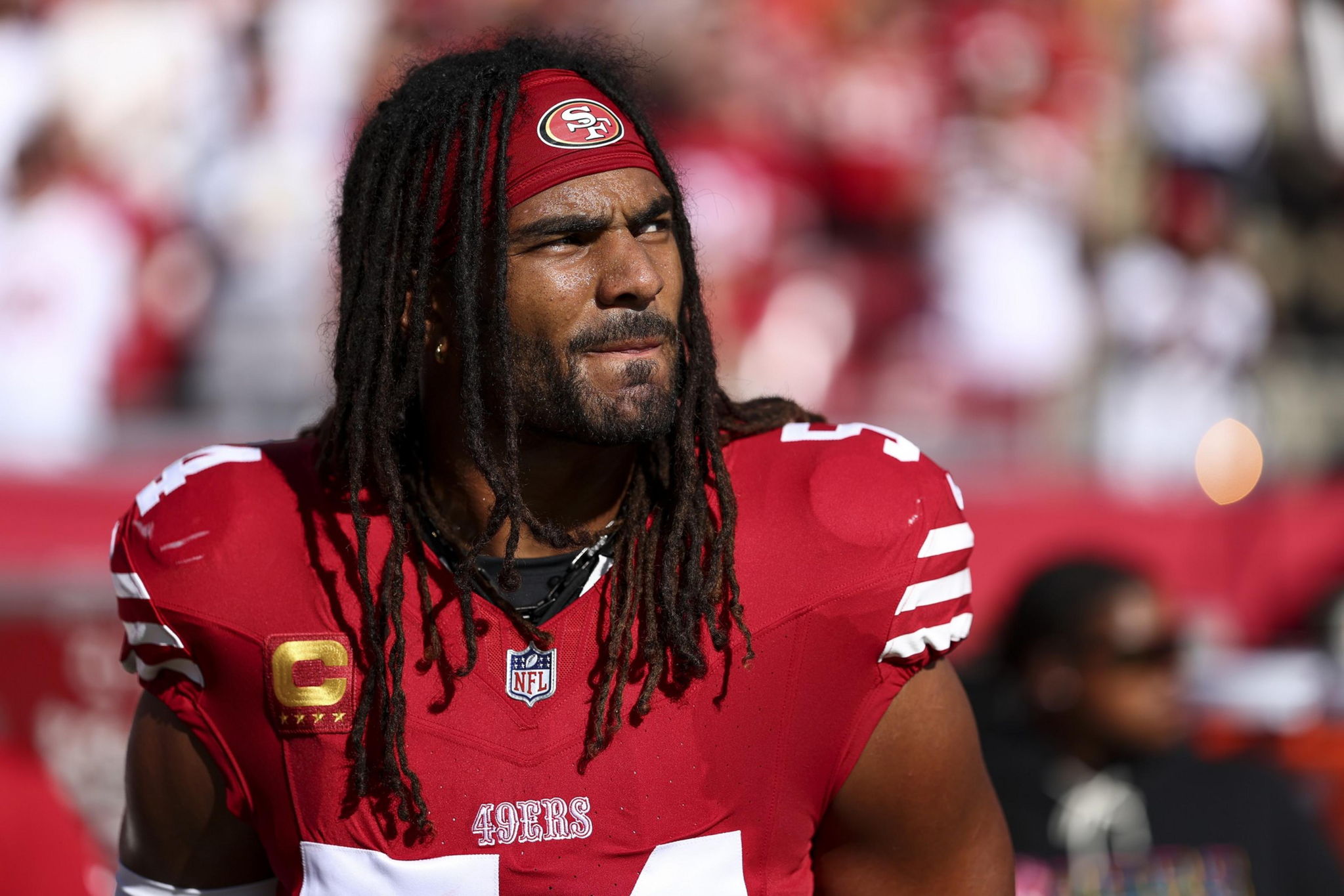 A football player with long dreadlocks wears a red San Francisco 49ers jersey and headband, looking off into the distance with a focused expression.