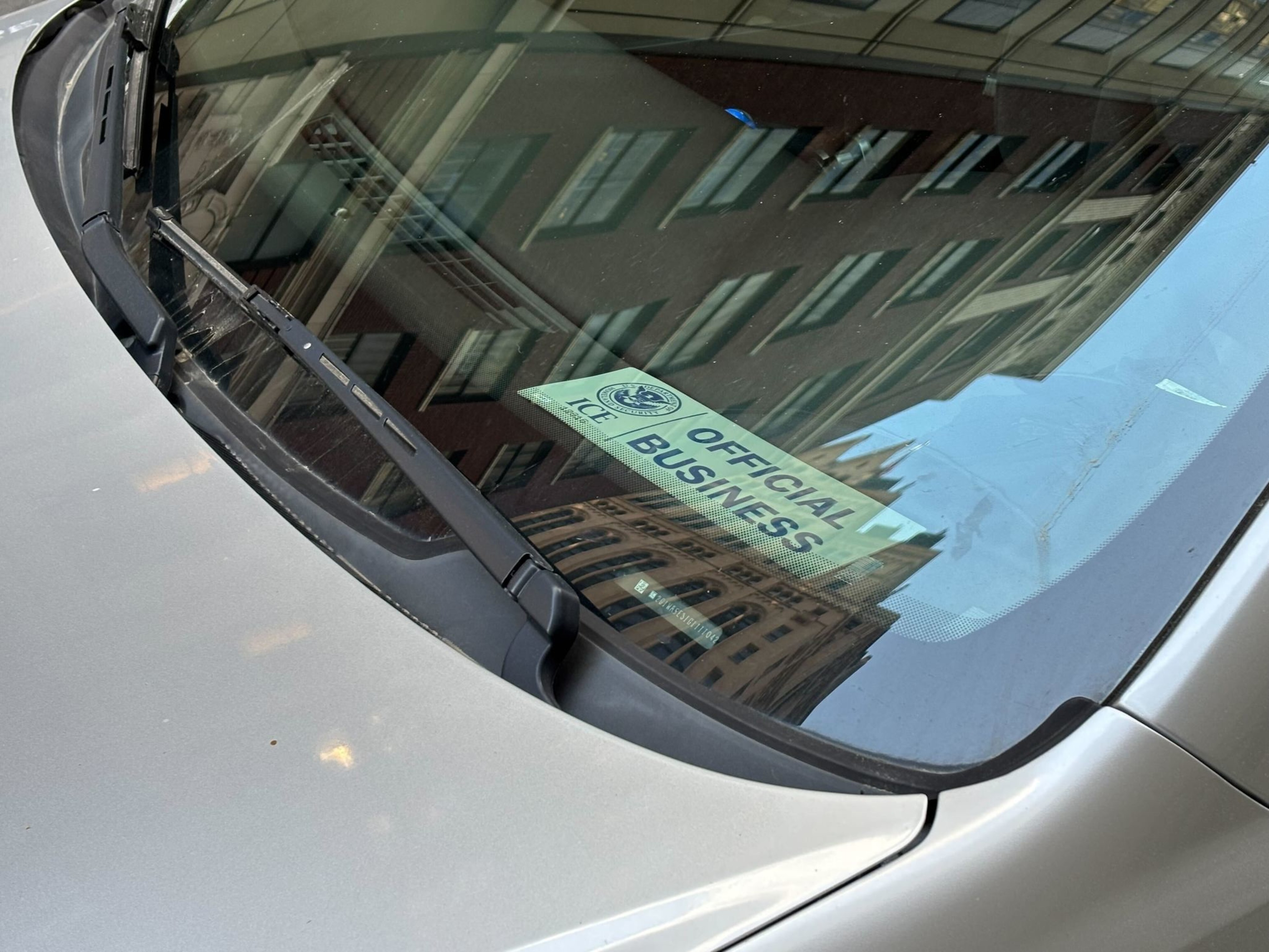 A car windshield displays a sign that reads &quot;OFFICIAL BUSINESS&quot; under a seal, with buildings reflecting off the glass.