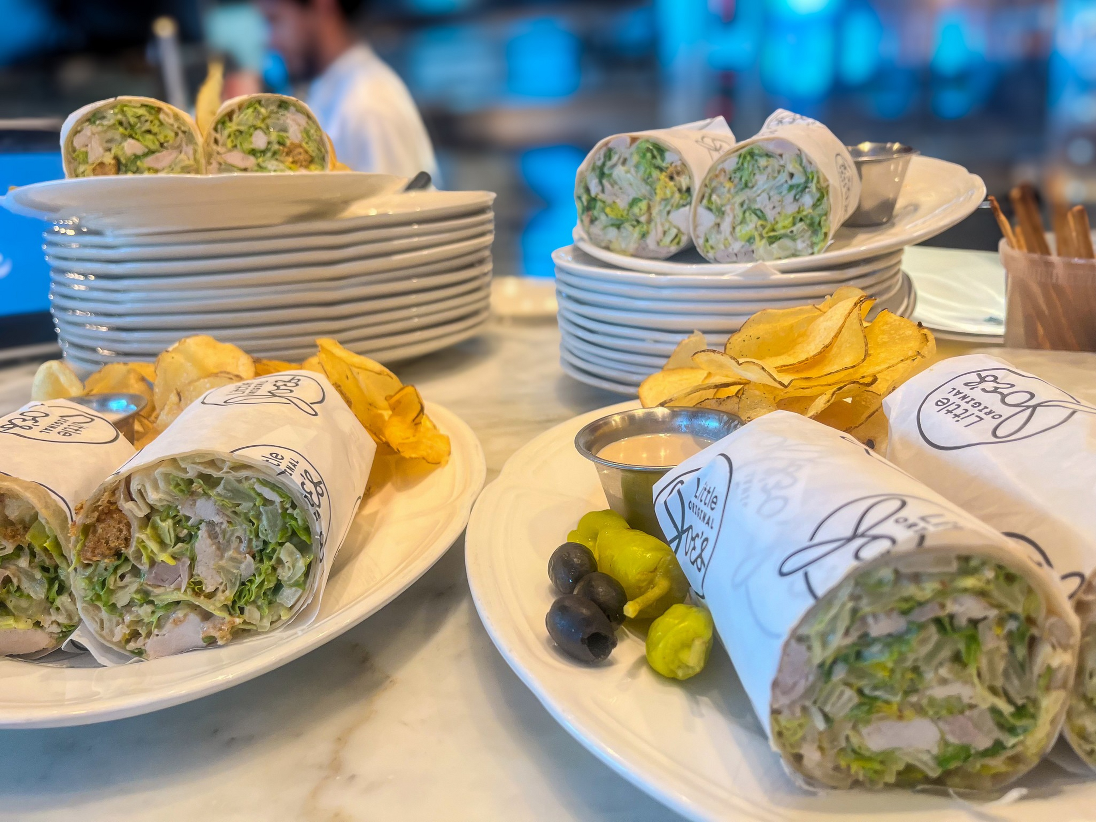 Chicken wraps filled with lettuce are cut in half and served on plates with potato chips, olives, pepperoncini, and dipping sauce.