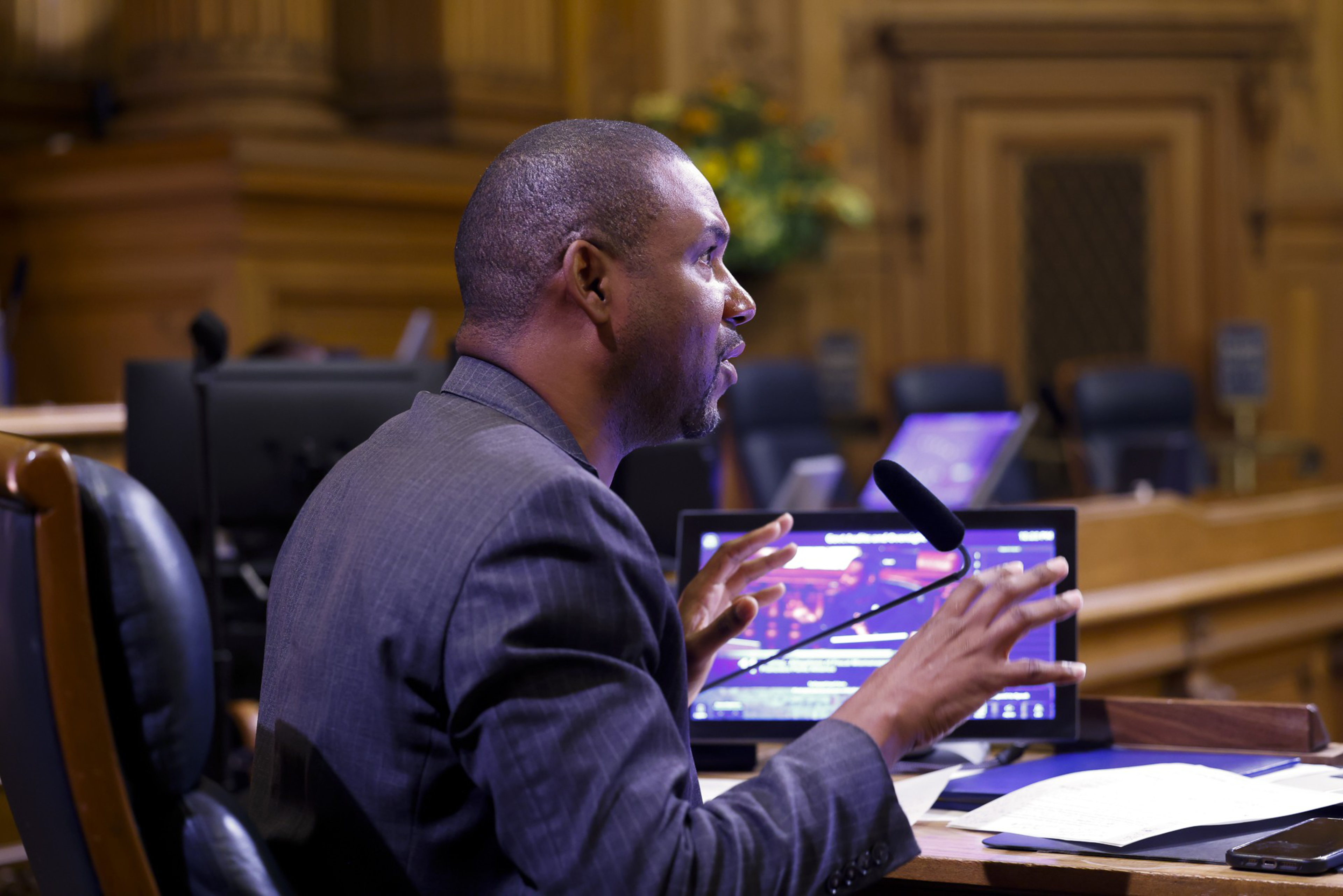 A man in a suit speaks passionately at a microphone in an ornate room. He gestures with his hands near a laptop and papers on a desk.