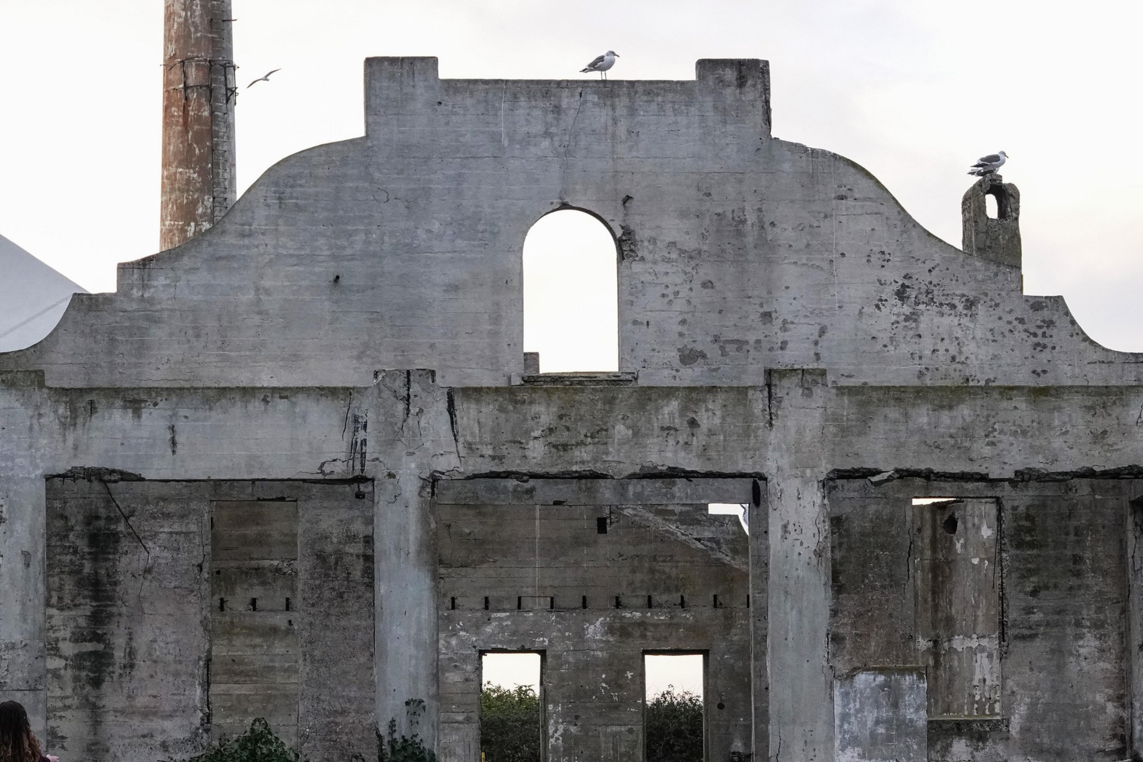 A weathered concrete structure with an arched opening at the top, accompanied by a few seagulls perched on it, and a tall chimney in the background.
