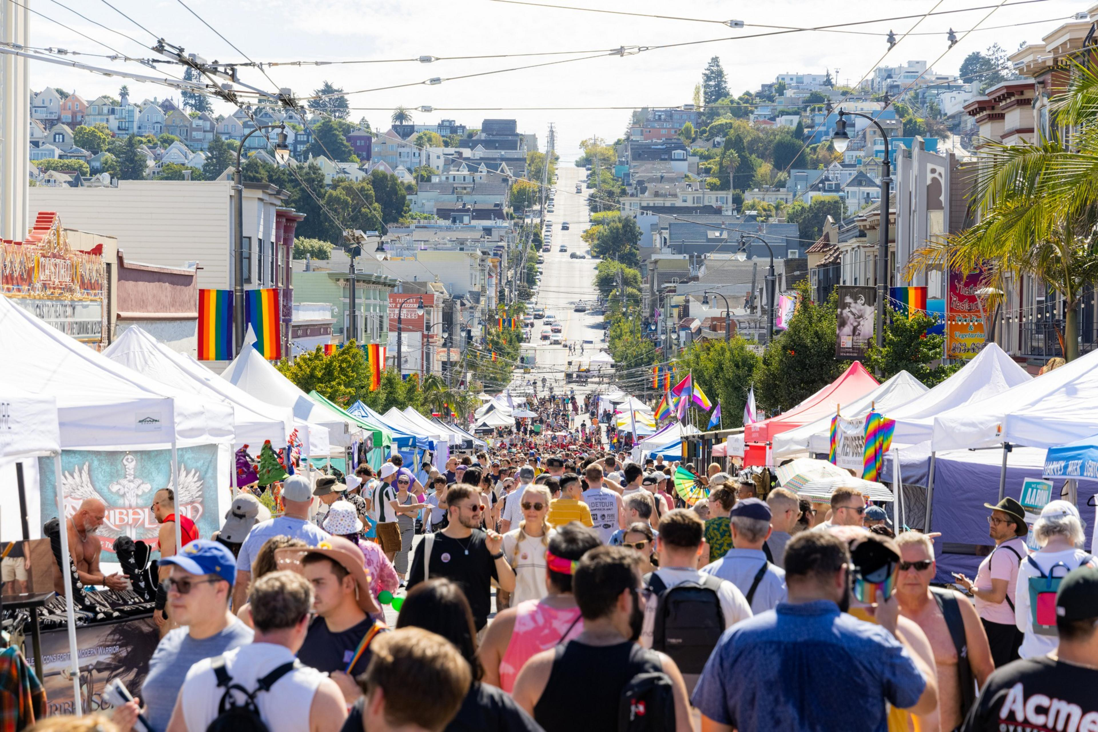 A bustling street festival with colorful tents, diverse crowds, and rainbow flags, set against a hilly background with houses and trees.