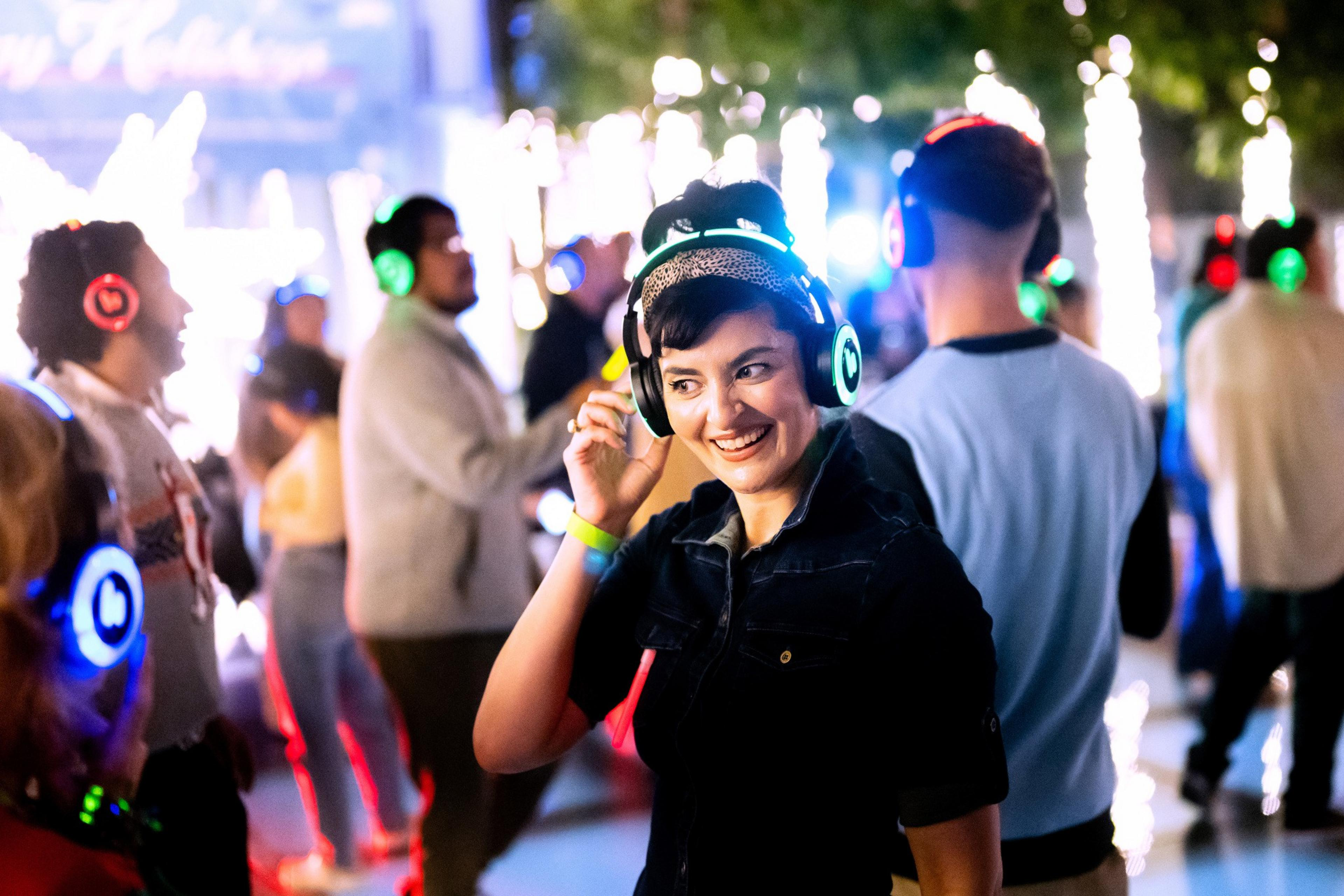 A lively scene shows people at a silent disco, wearing glowing headphones in various colors. A smiling woman in the foreground adjusts hers while dancing.