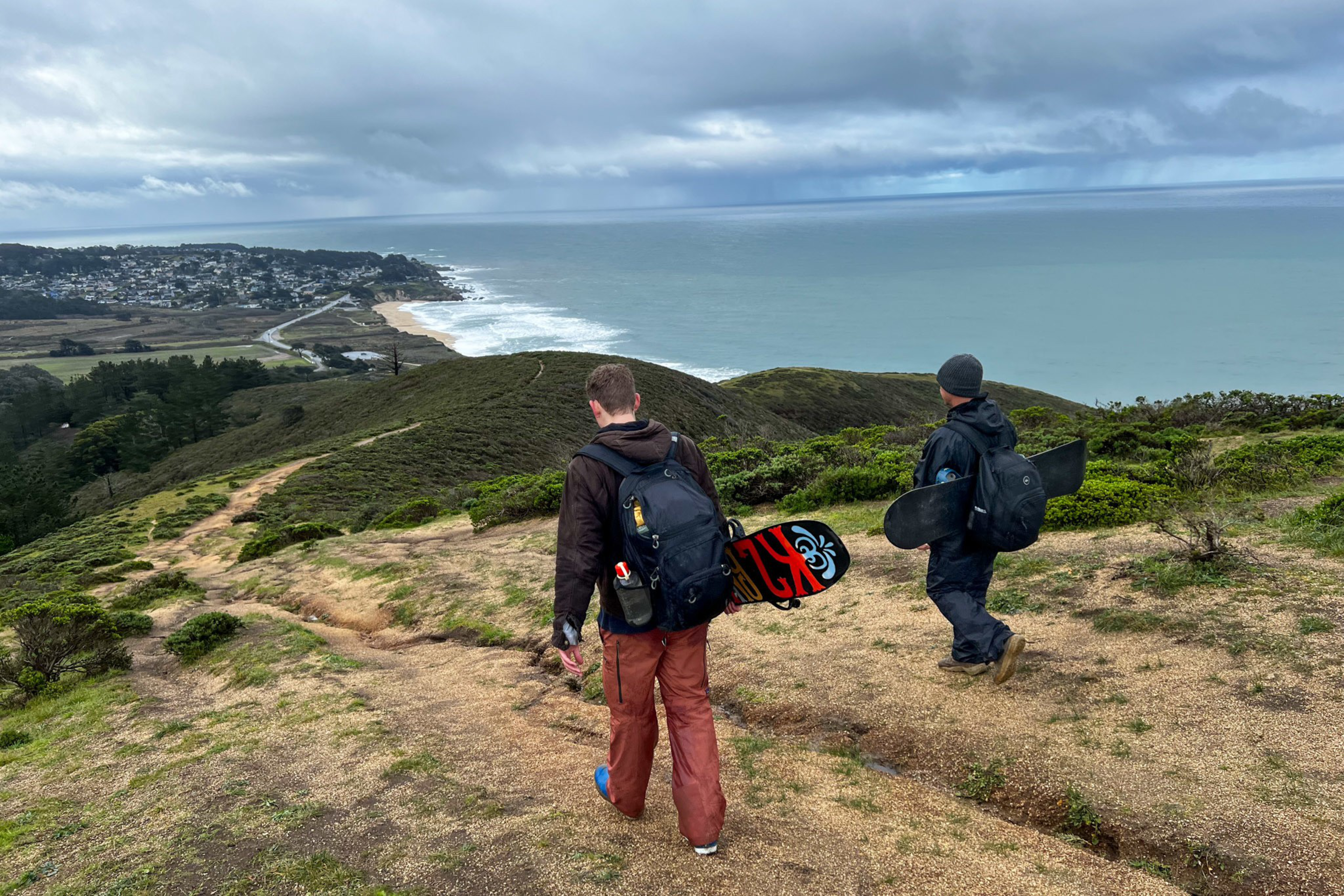 Two people walk down a dirt path on a grassy hillside toward the ocean, each carrying a longboard with cloudy skies overhead.