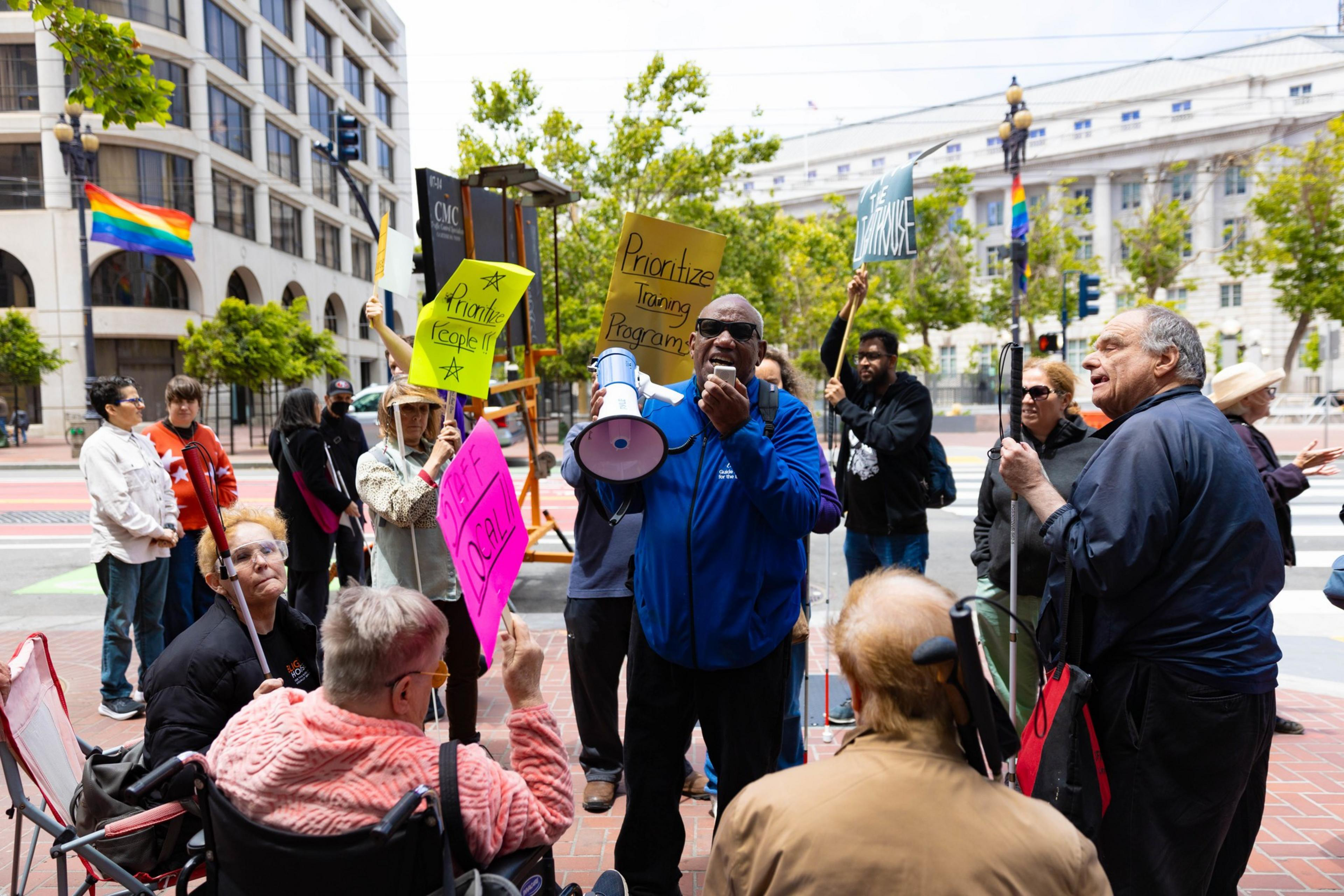 People gather on a city street holding signs calling for prioritizing people and training programs. A man speaks into a megaphone amid a diverse crowd.
