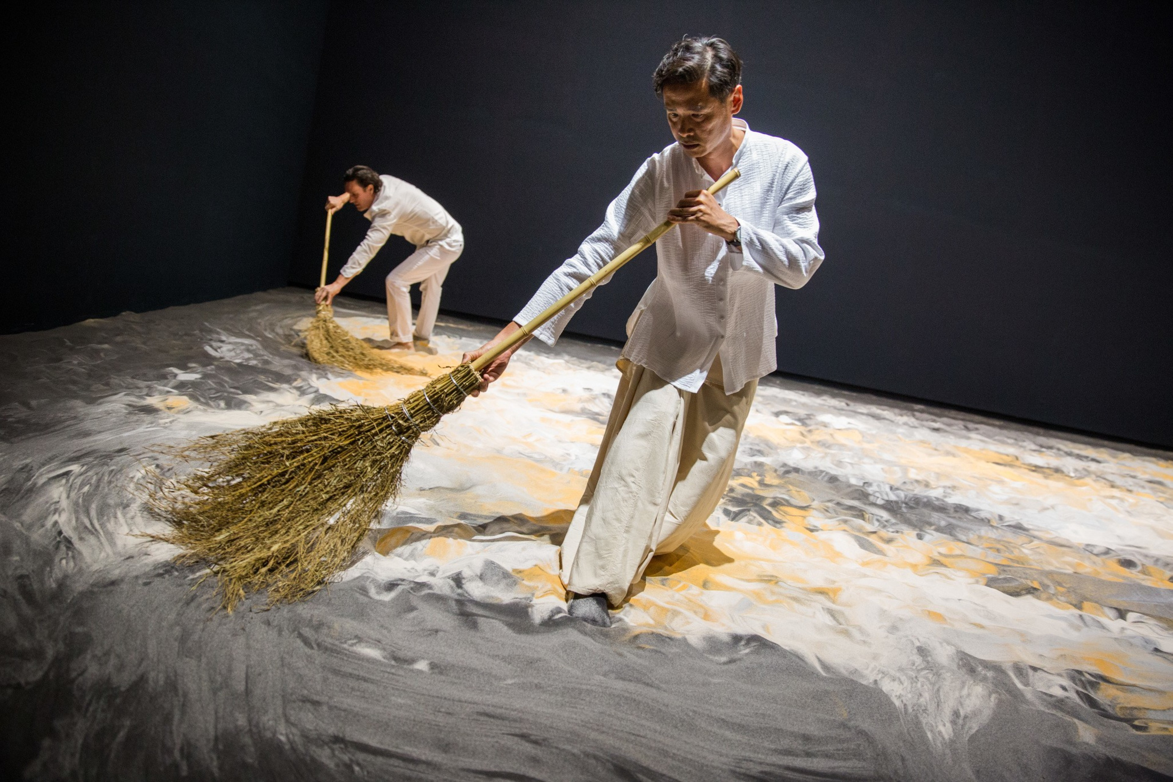 Two people sweep a sandy floor with large brooms in a dark room, creating a textured pattern.