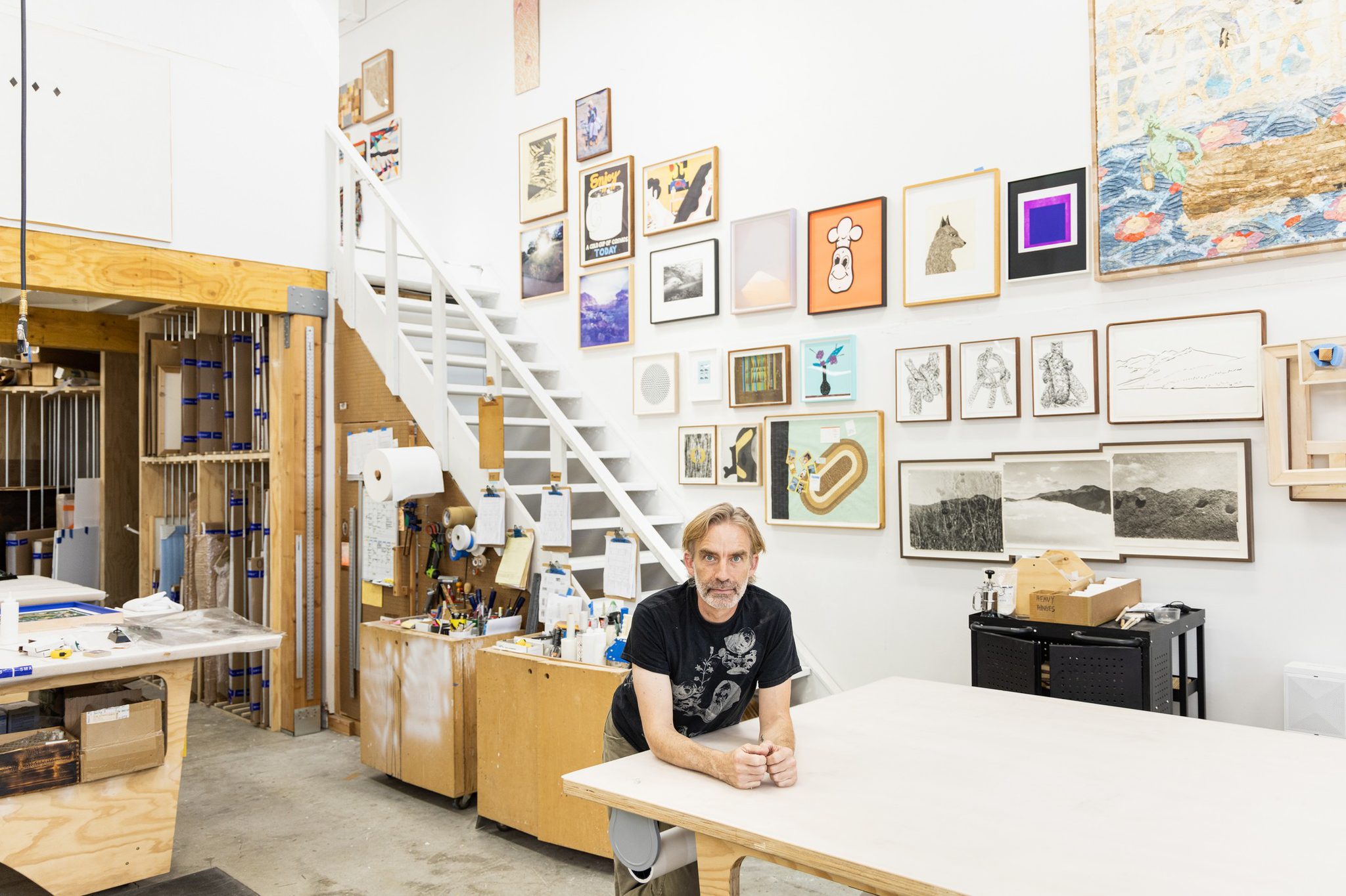 A man with gray hair and a beard leans on a large white table in a bright studio filled with wooden furniture and colorful artwork on the walls.